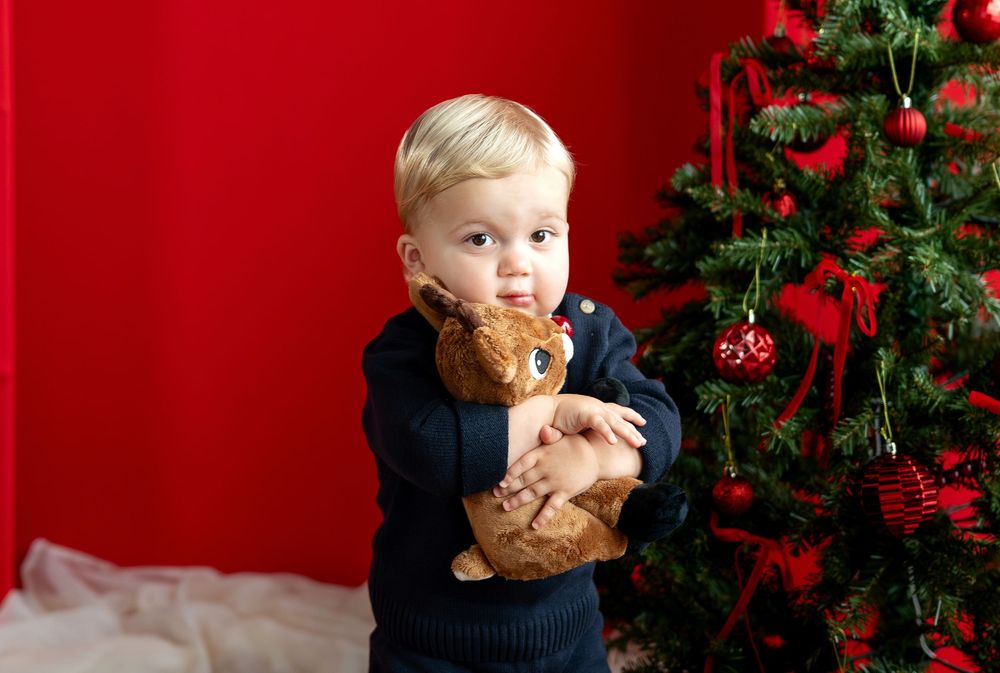 Cute toddler in navy sweater hugs stuffed reindeer toy beside Christmas tree.