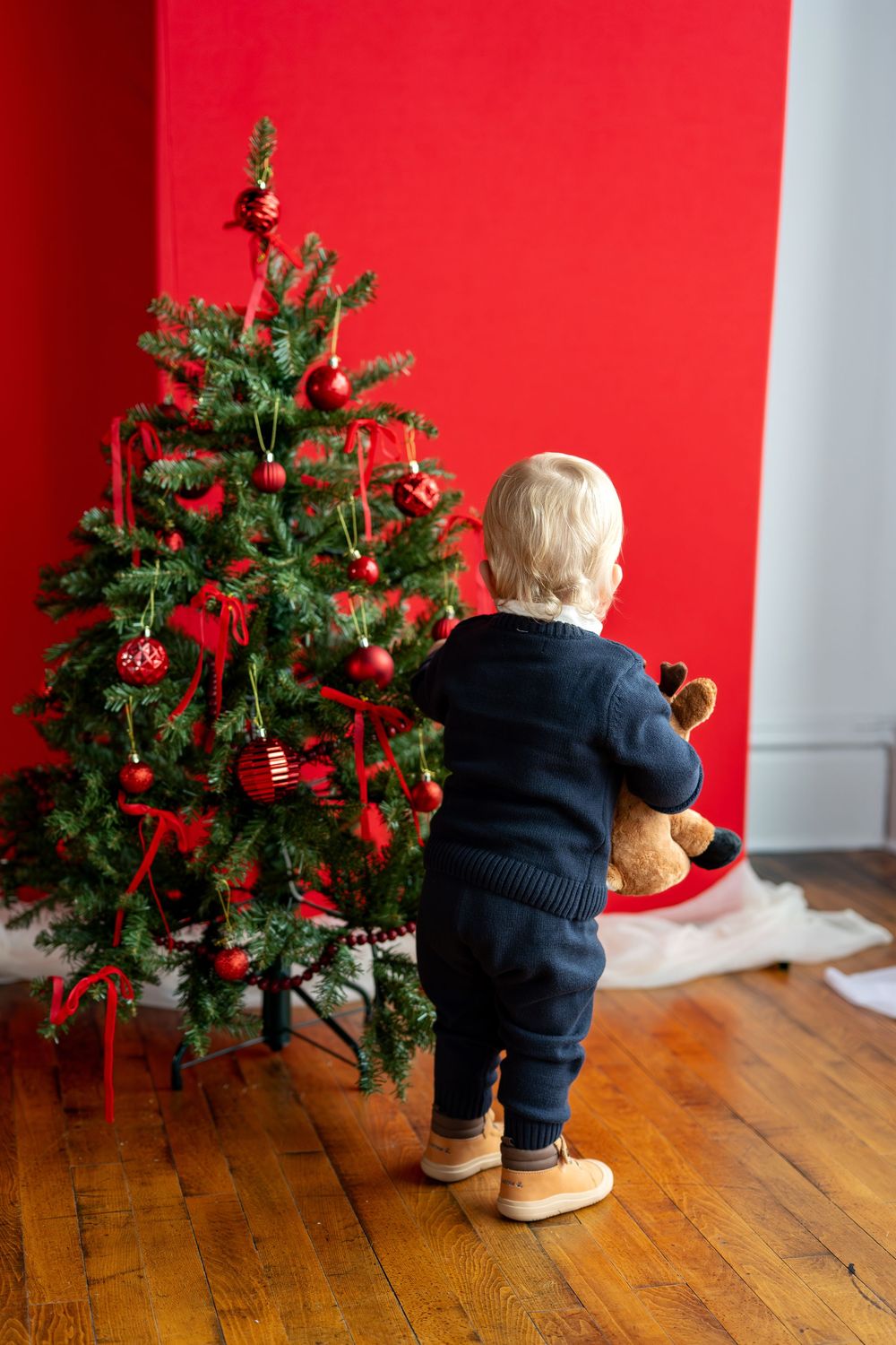 Toddler in navy outfit holds stuffed reindeer while exploring decorated Christmas tree.