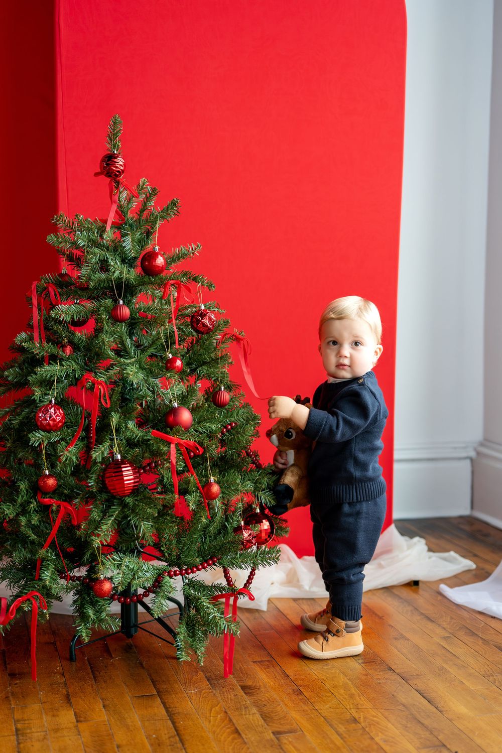 Toddler in navy outfit holds stuffed reindeer beside beautifully decorated Christmas tree.