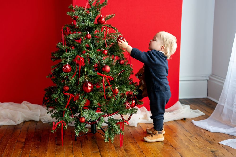 Toddler in navy outfit decorating a Christmas tree with red ornaments against a bold red wall.