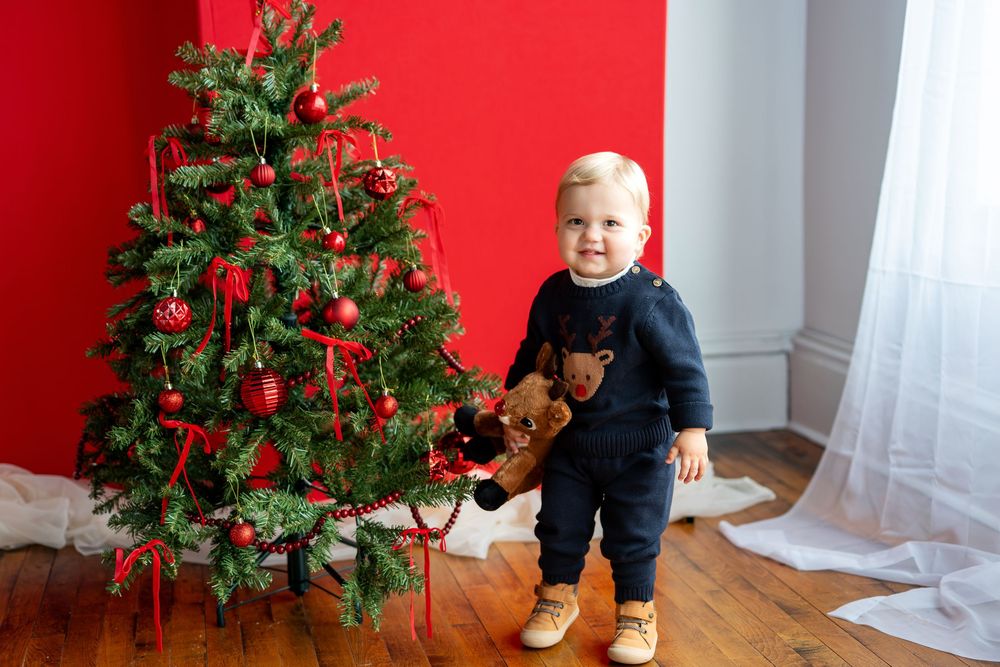 Happy toddler in navy reindeer sweater stands with stuffed toy by Christmas tree.
