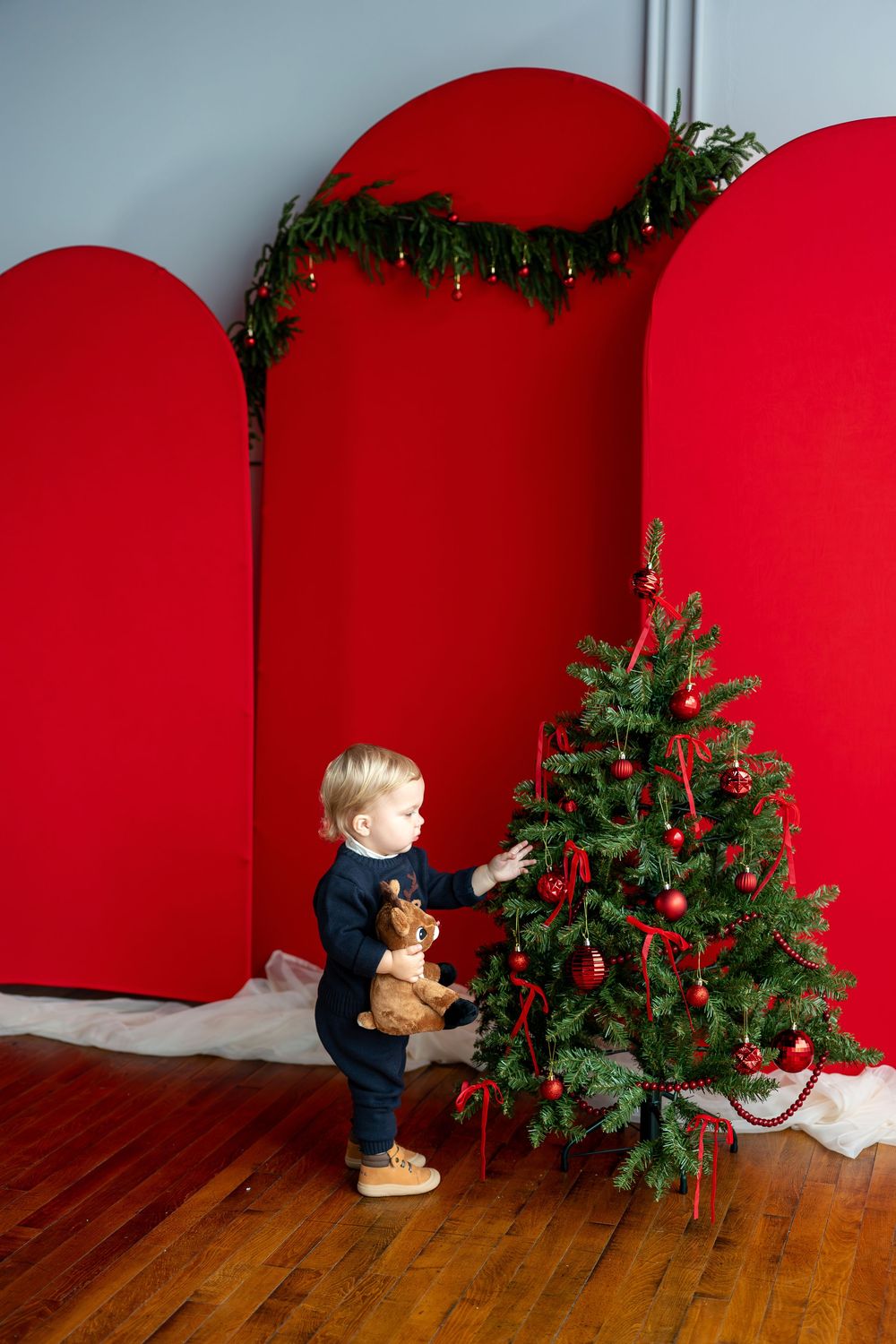 Smiling toddler holding a reindeer stuffed animal beside a decorated Christmas tree with red backdrop.