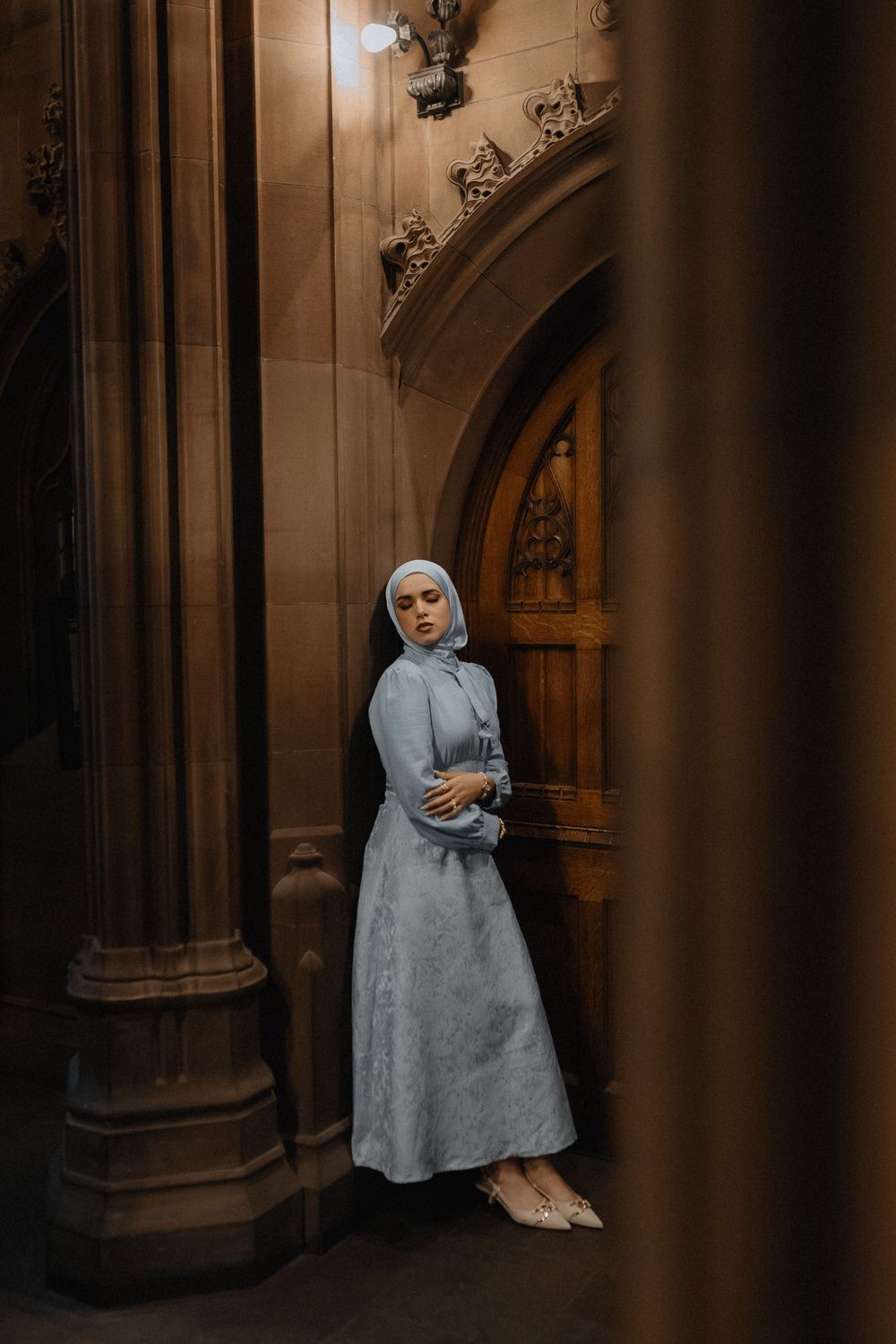 A figure in a light blue dress stands in an ornate Gothic archway with dramatic shadows and architectural details.