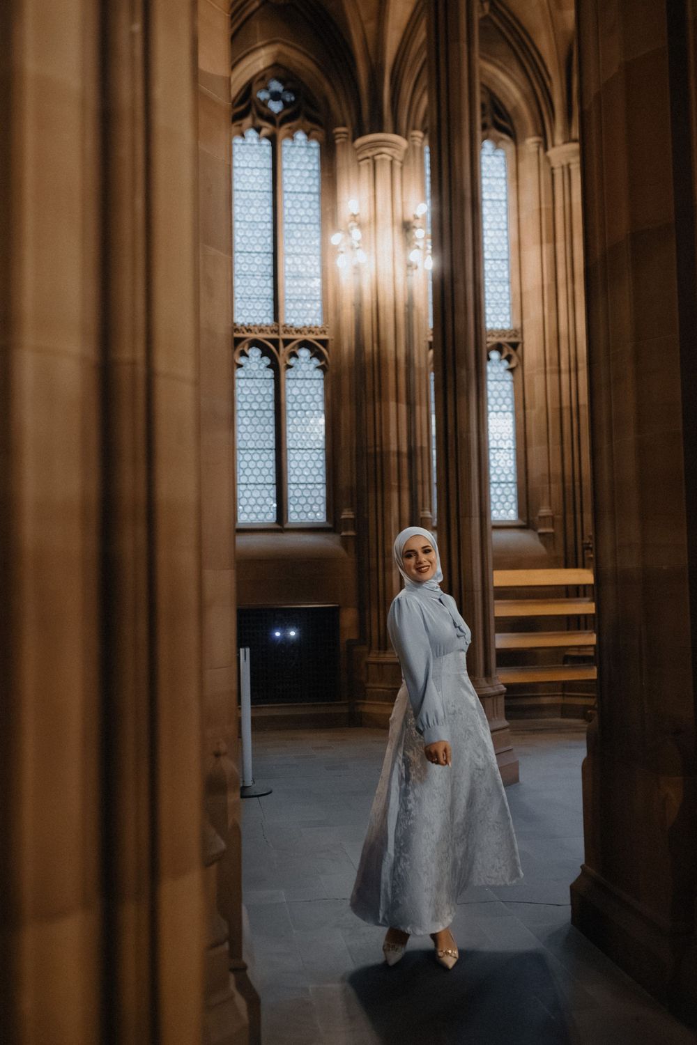 Person in flowing grey dress moves gracefully through ornate cathedral hallway with arched windows.