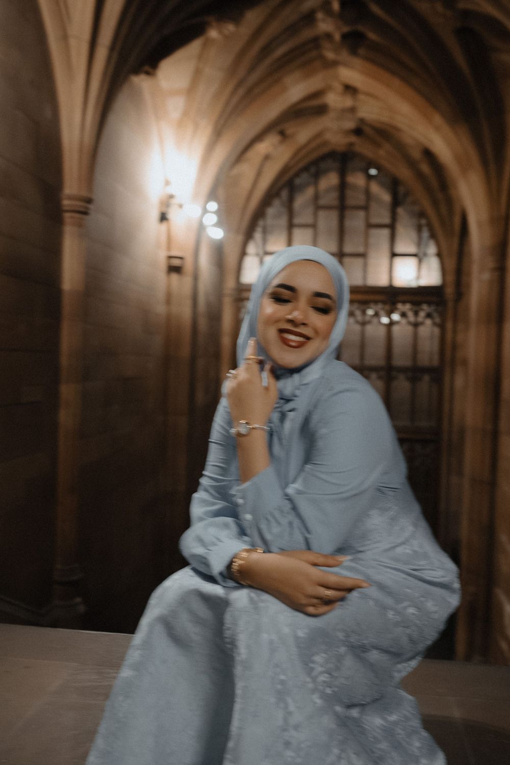 A person in a light blue hijab and dress laughs joyfully while standing in an ornate stone archway corridor.