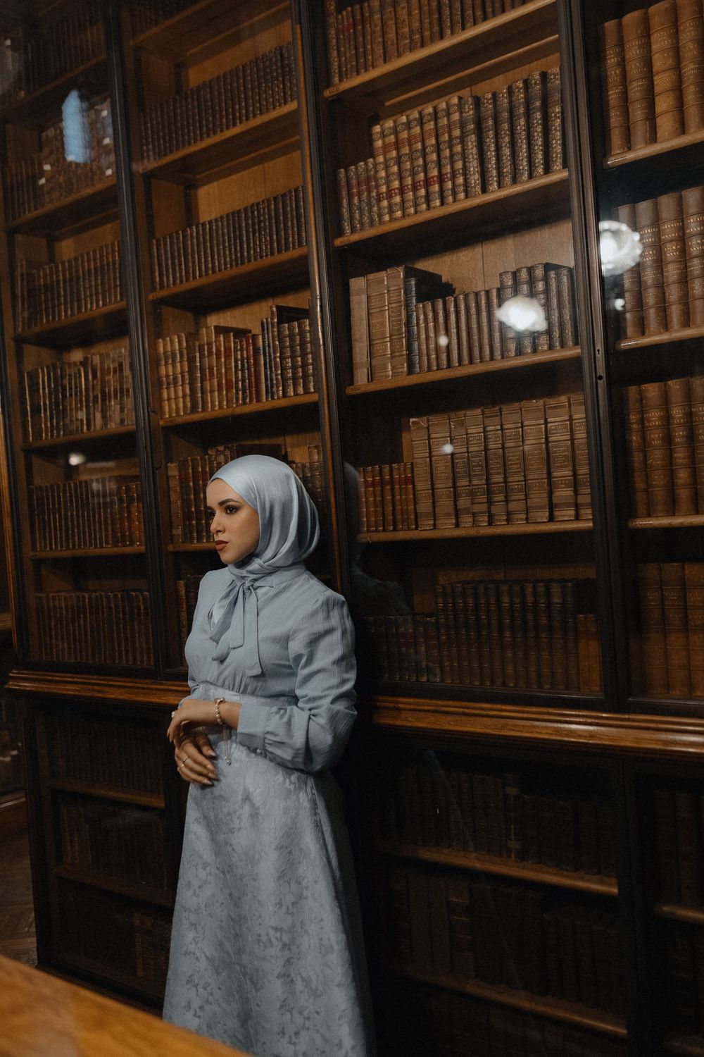 A person poses against vintage bookshelves in a historic library wearing a light blue ensemble.