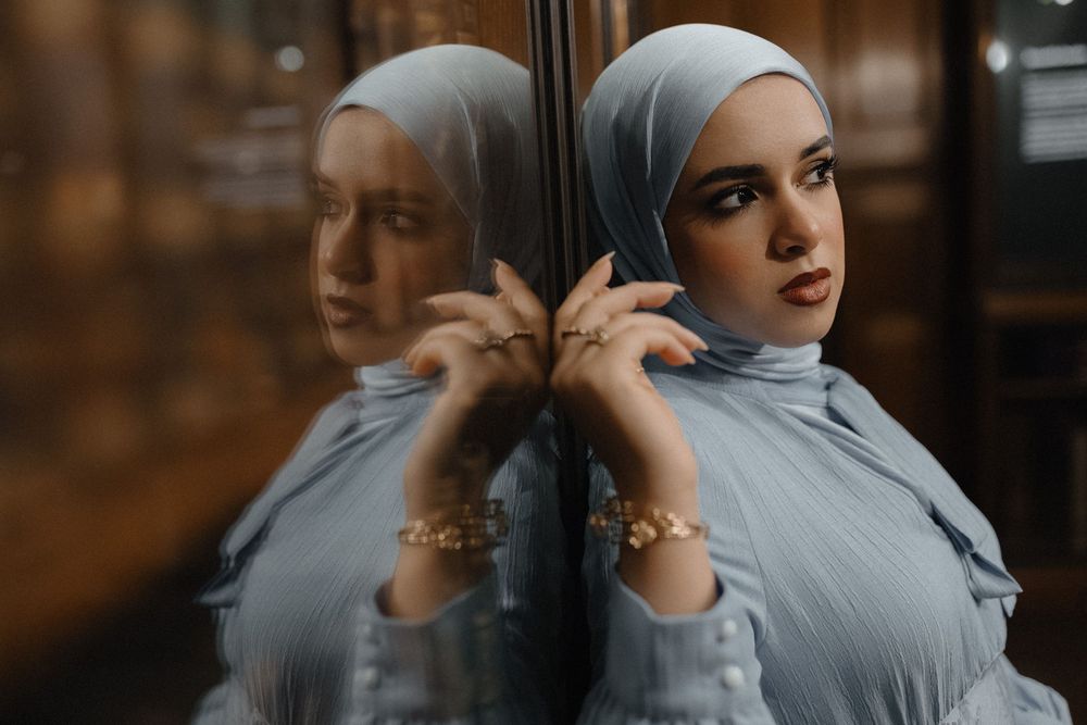 A person in a grey hijab poses against a reflective surface creating a dramatic mirrored effect in moody lighting.