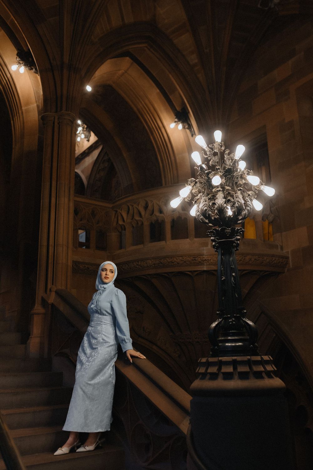 Ornate gothic architecture interior with stone arches and vintage lamp post illuminating a figure in light blue attire.