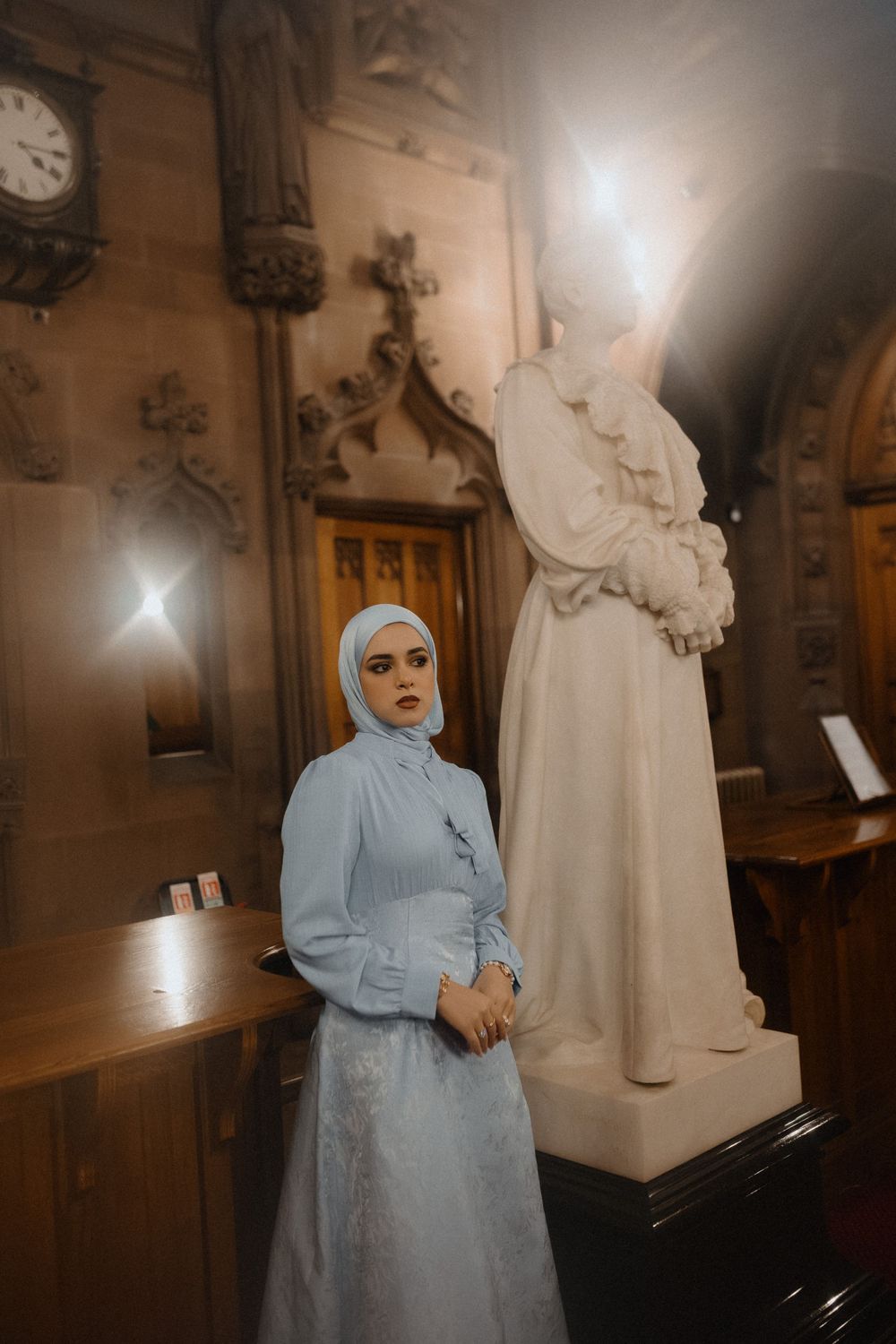 A person in a light blue hijab and dress stands near a piano in a dimly lit church interior with ornate architectural details.