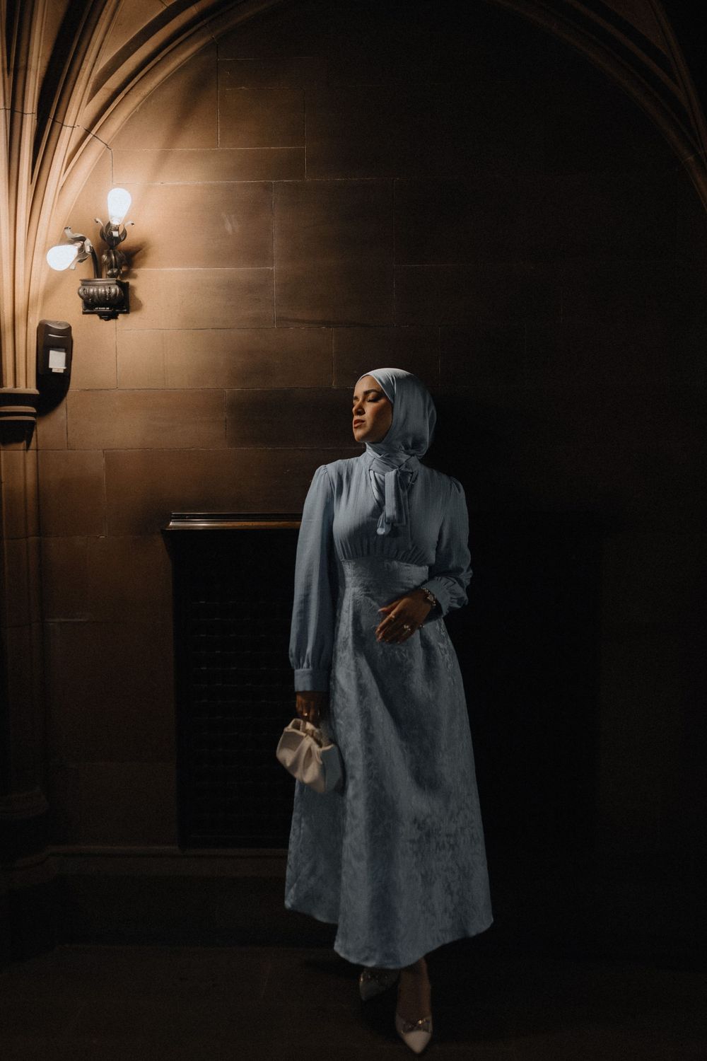 Figure in grey cloak walks through dimly lit gothic corridor with dramatic lighting and shadows.