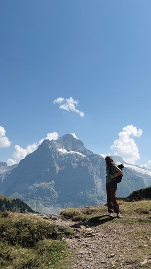 Hiker with backpack standing on a mountain trail admiring a snow-capped Alpine peak under a blue sky.
