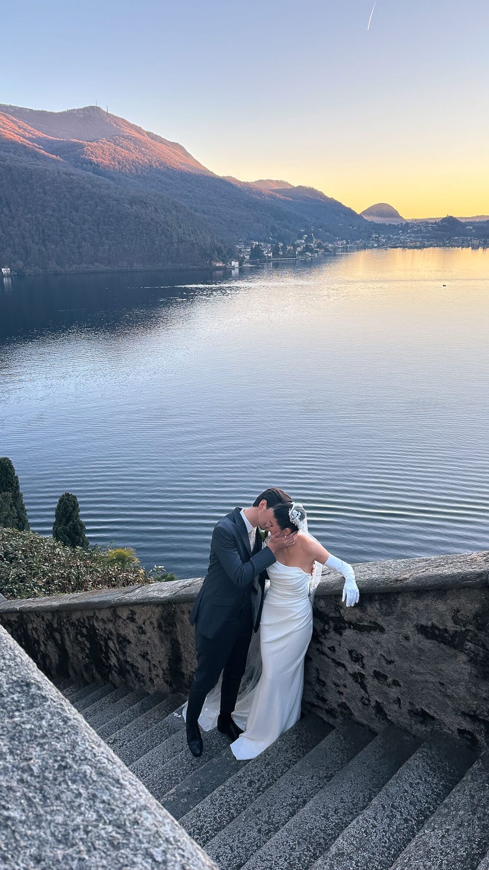 Bride and groom kissing on stone steps by Lake Como at sunset with mountain reflections on calm water.