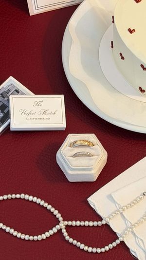 Wedding rings in velvet box beside pearl necklace, cake, and The Perfect Match card on red table.