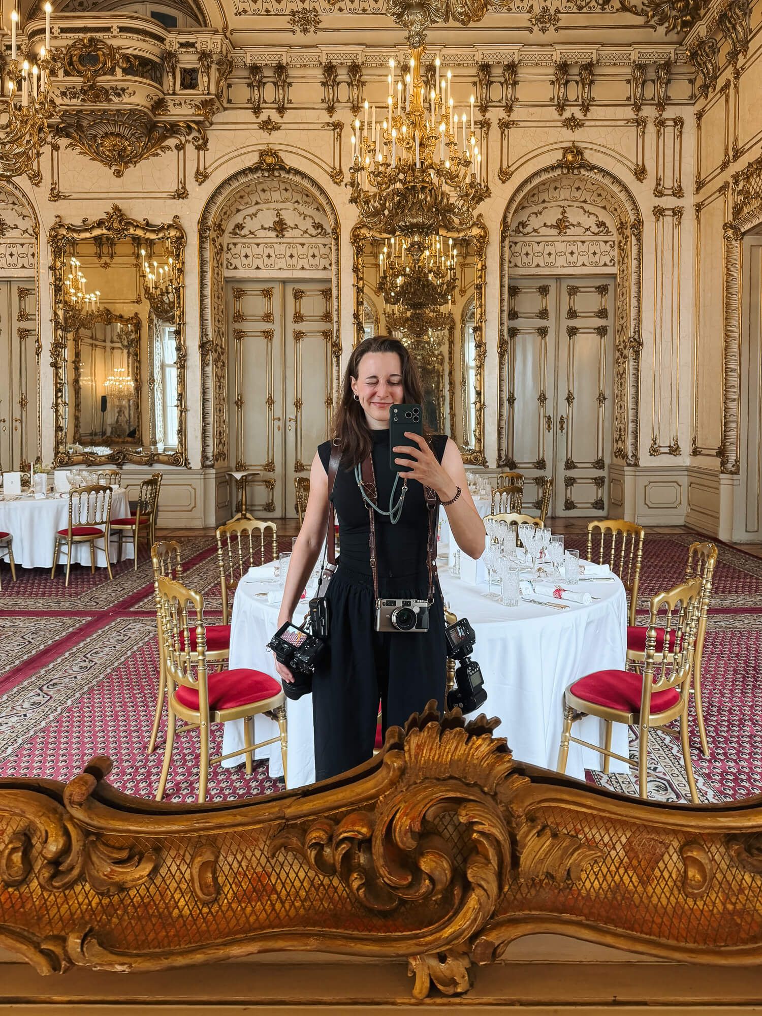 Woman in black dress takes mirror selfie in ornate gilded ballroom with chandeliers, red carpet, and elegant dining tables.