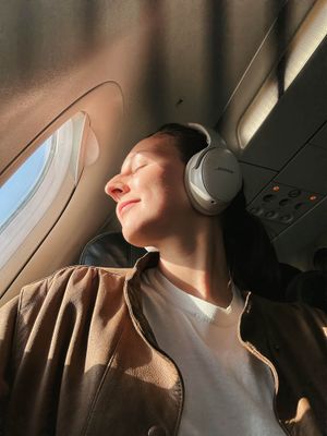 Person wearing Bose headphones relaxing in airplane seat with eyes closed, bathed in warm sunlight from window.