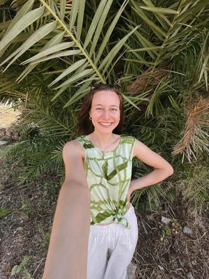 Young woman smiling in green tropical print top standing among lush palm trees outdoors on a sunny day.