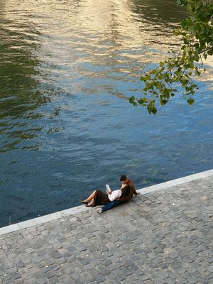 Two people sitting on a cobblestone riverbank beside calm blue water, relaxing with a laptop on a sunny day.