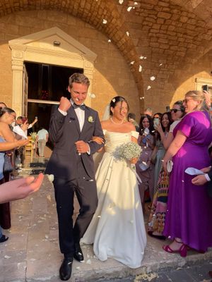 Newlywed couple exits stone church amid confetti, bride in white gown with bouquet, groom in black tuxedo, guests cheering.