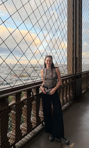 Woman with camera poses on Eiffel Tower observation deck with Paris cityscape and diamond grid fence behind her.