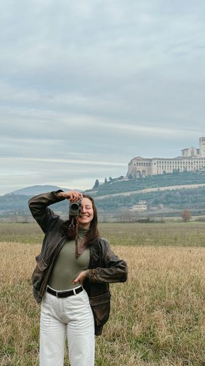 Person in olive green sweater and white jeans poses in field with historic hilltop monastery of Assisi in background.