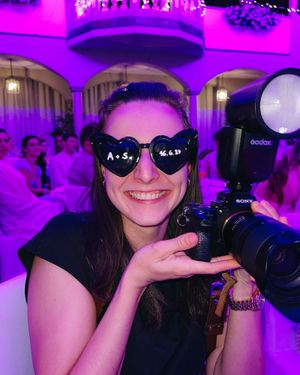 A photographer in sunglasses poses playfully with their camera equipment in a purple-lit event space.