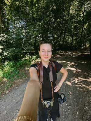 A person wearing a black jumpsuit and multiple cameras poses on a shaded forest trail on a sunny day.
