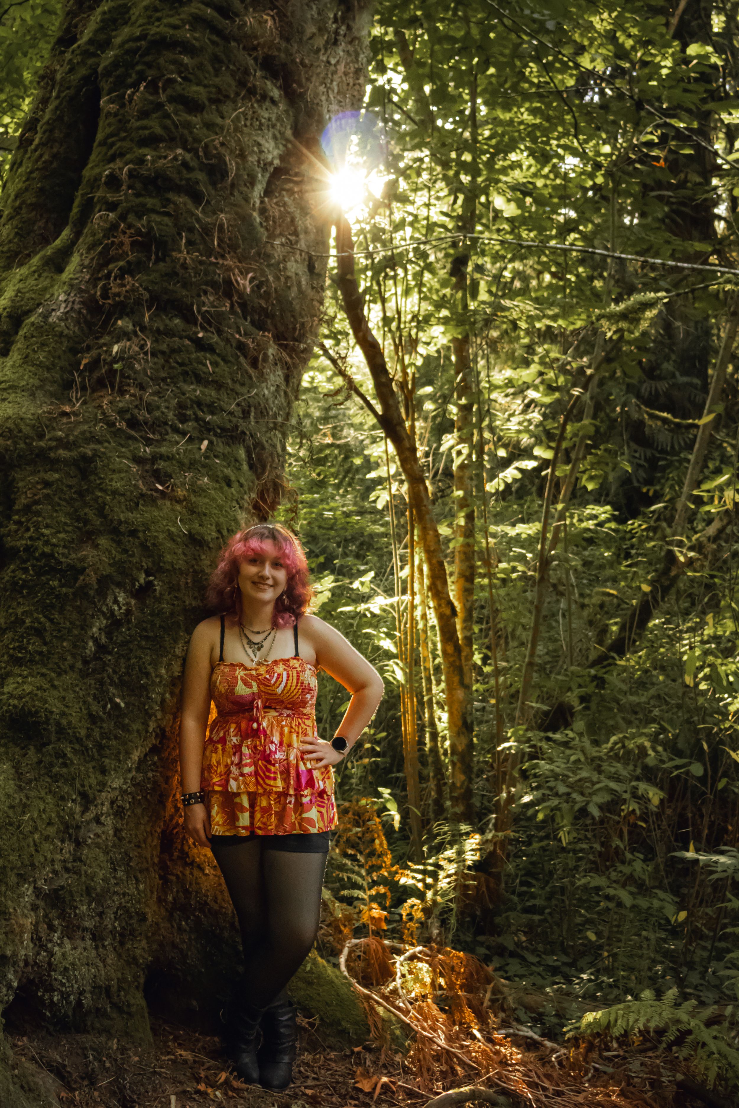Woman with pink hair in colorful dress posing by mossy tree in sunlit forest.