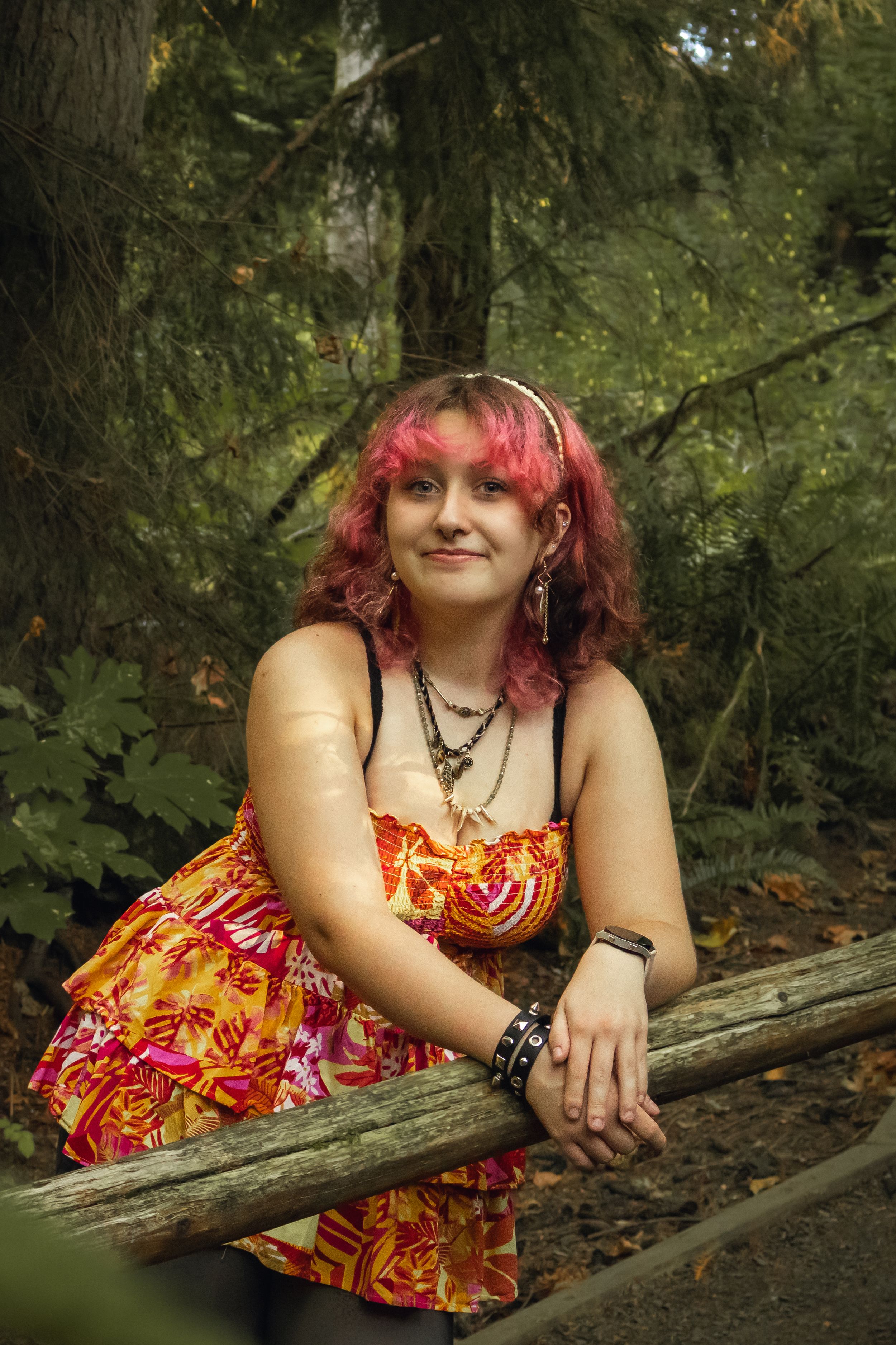 Smiling young woman with pink hair in a floral dress leaning on a wooden fence rail in a lush forest setting.