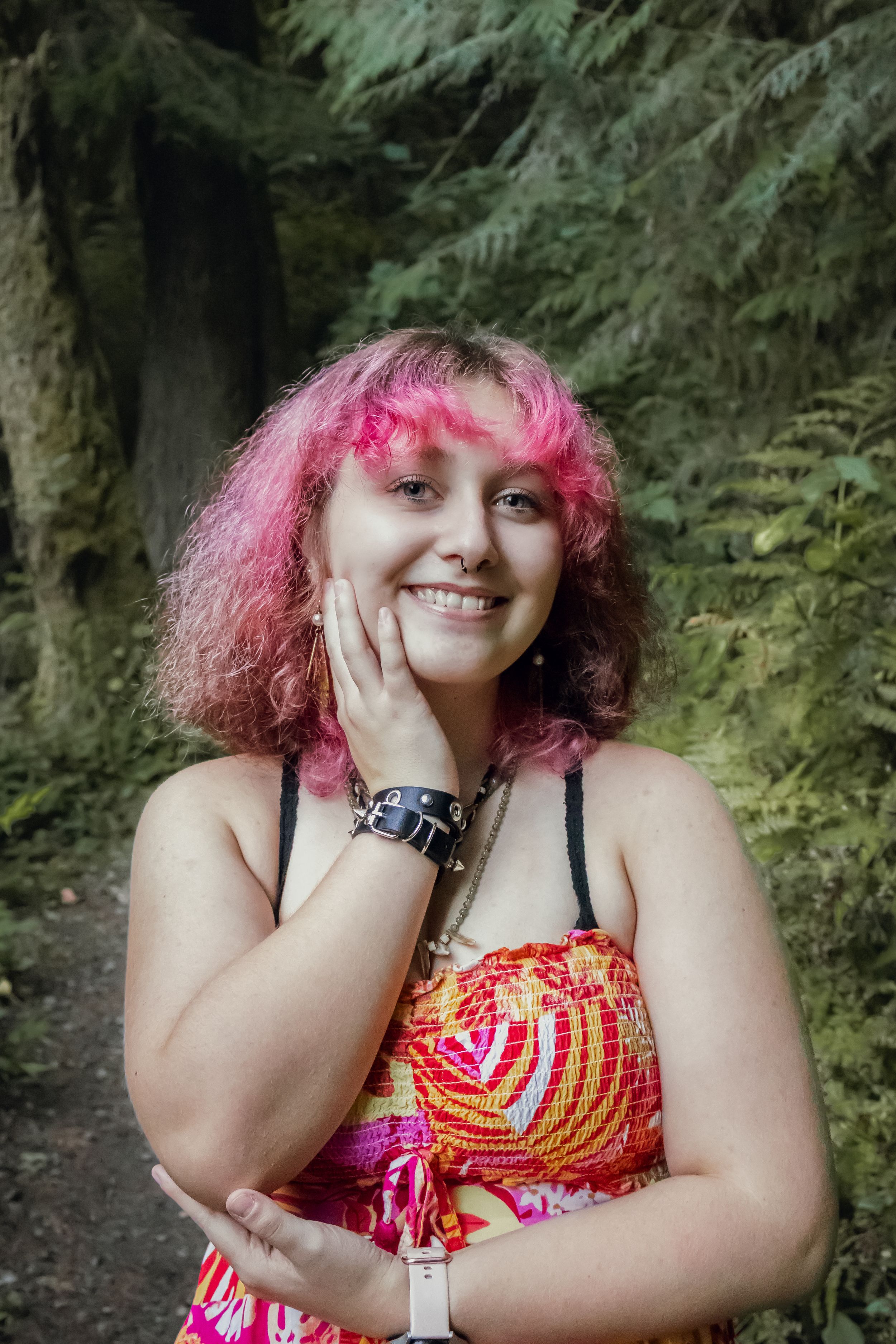 Smiling young woman with pink hair in a colorful dress touching her face with a spiked bracelet in a forest setting.