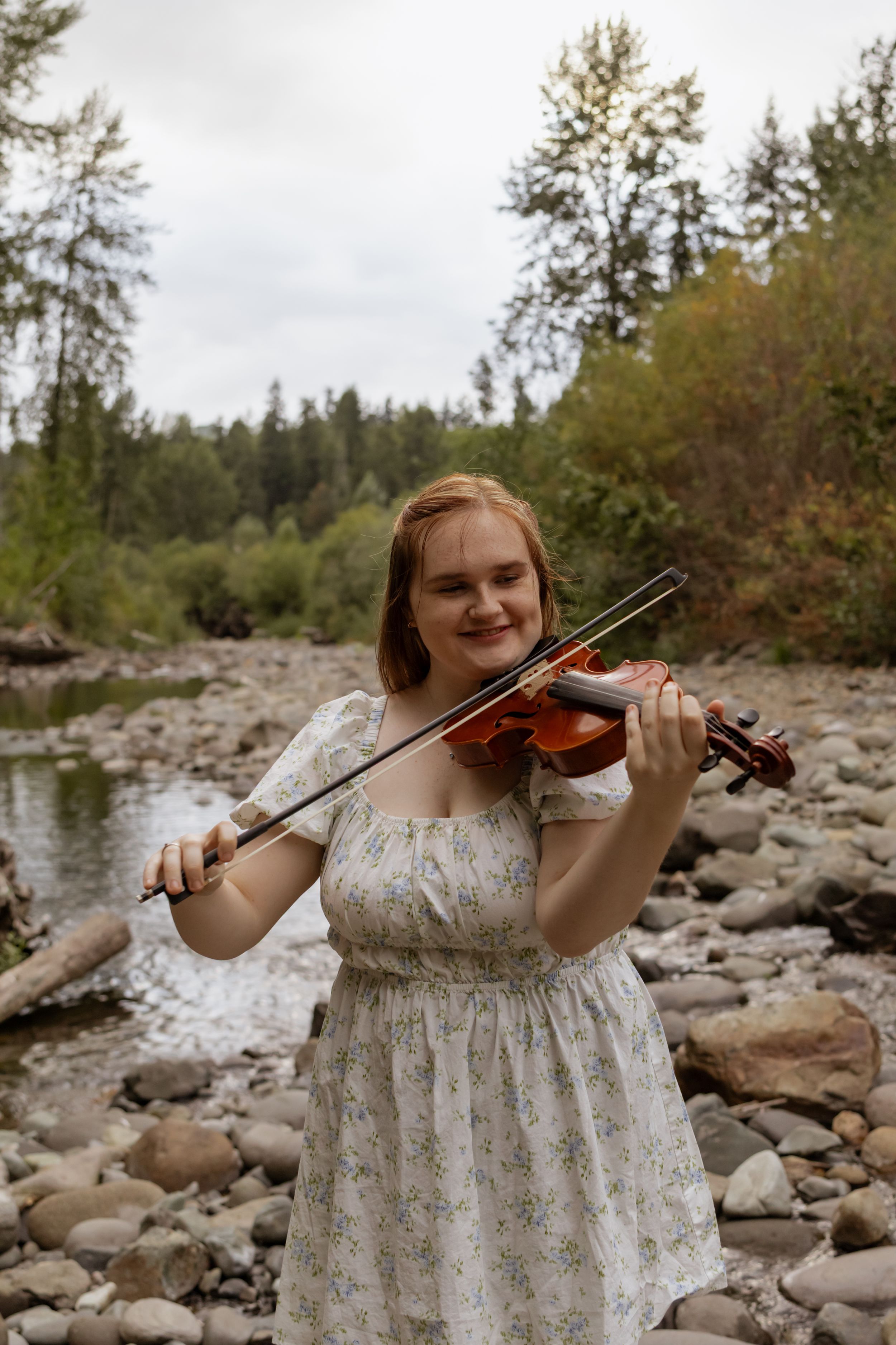 Young woman in floral dress playing violin outdoors by a rocky river with lush green forest background.