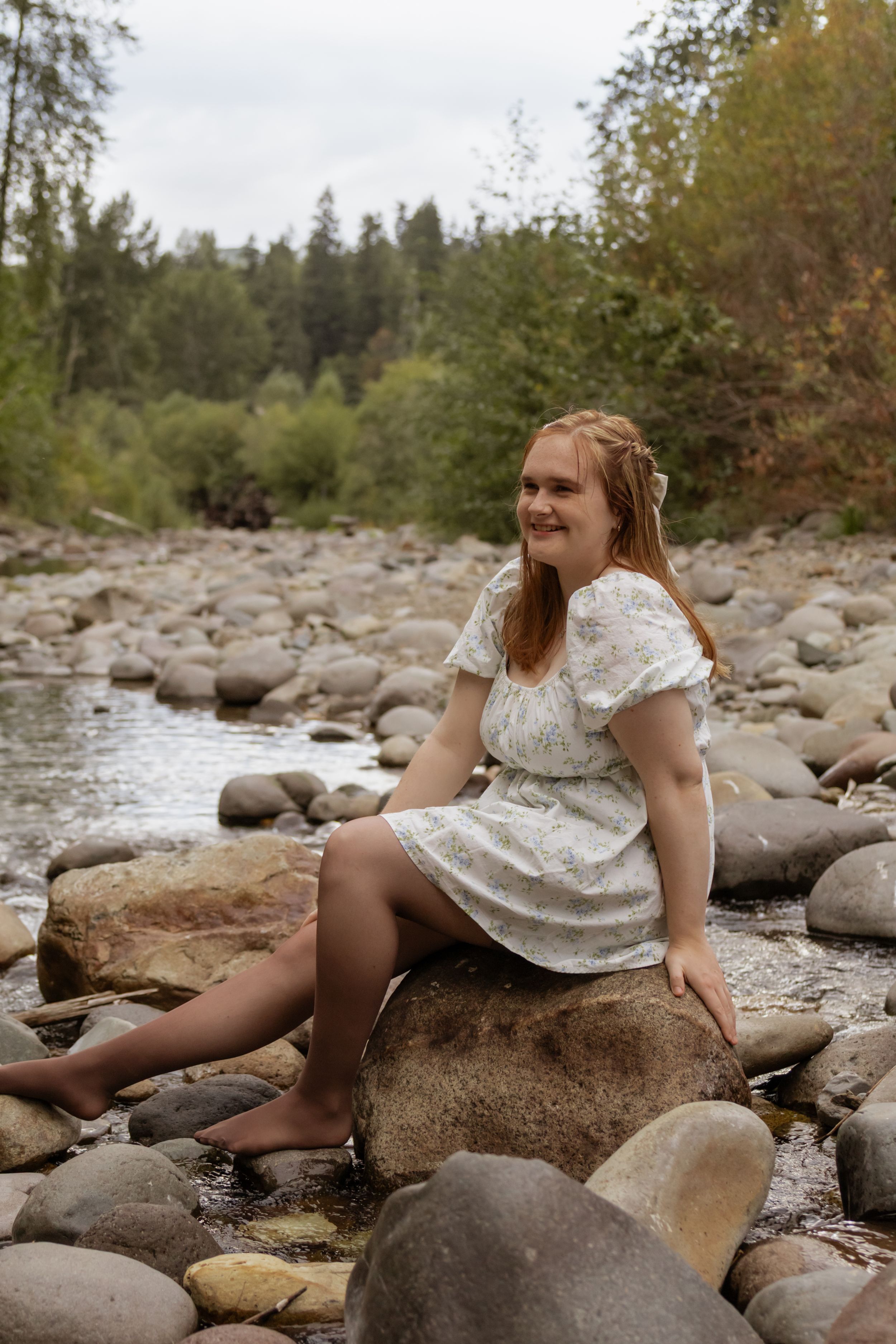 Young woman in white dress sitting on rocks by a peaceful forest river, smiling outdoors.