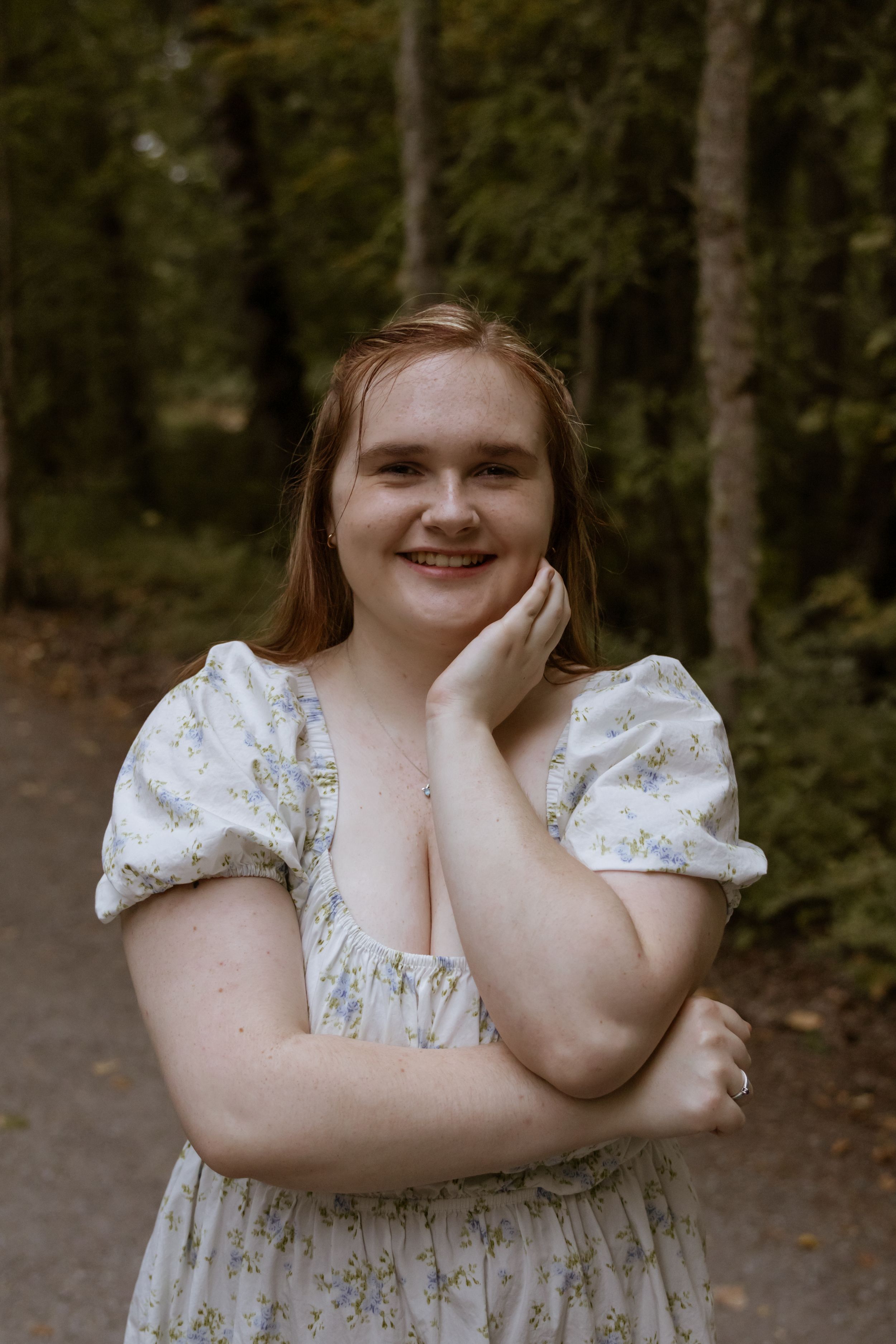 Young woman in floral white dress smiling in a forest path, posing with hand on cheek.