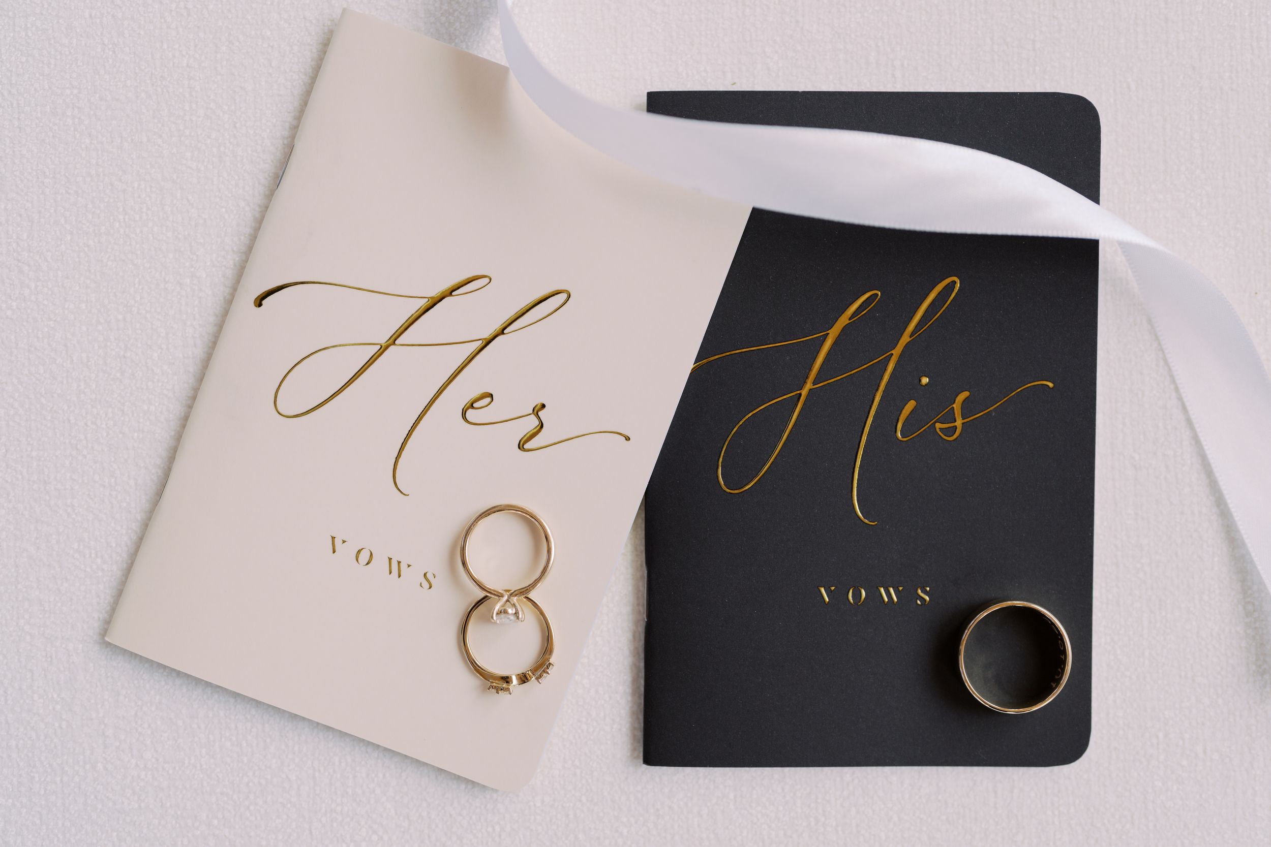 Wedding rings placed on elegant black and cream colored cards with gold calligraphy text reading 'His' and 'Hers'.