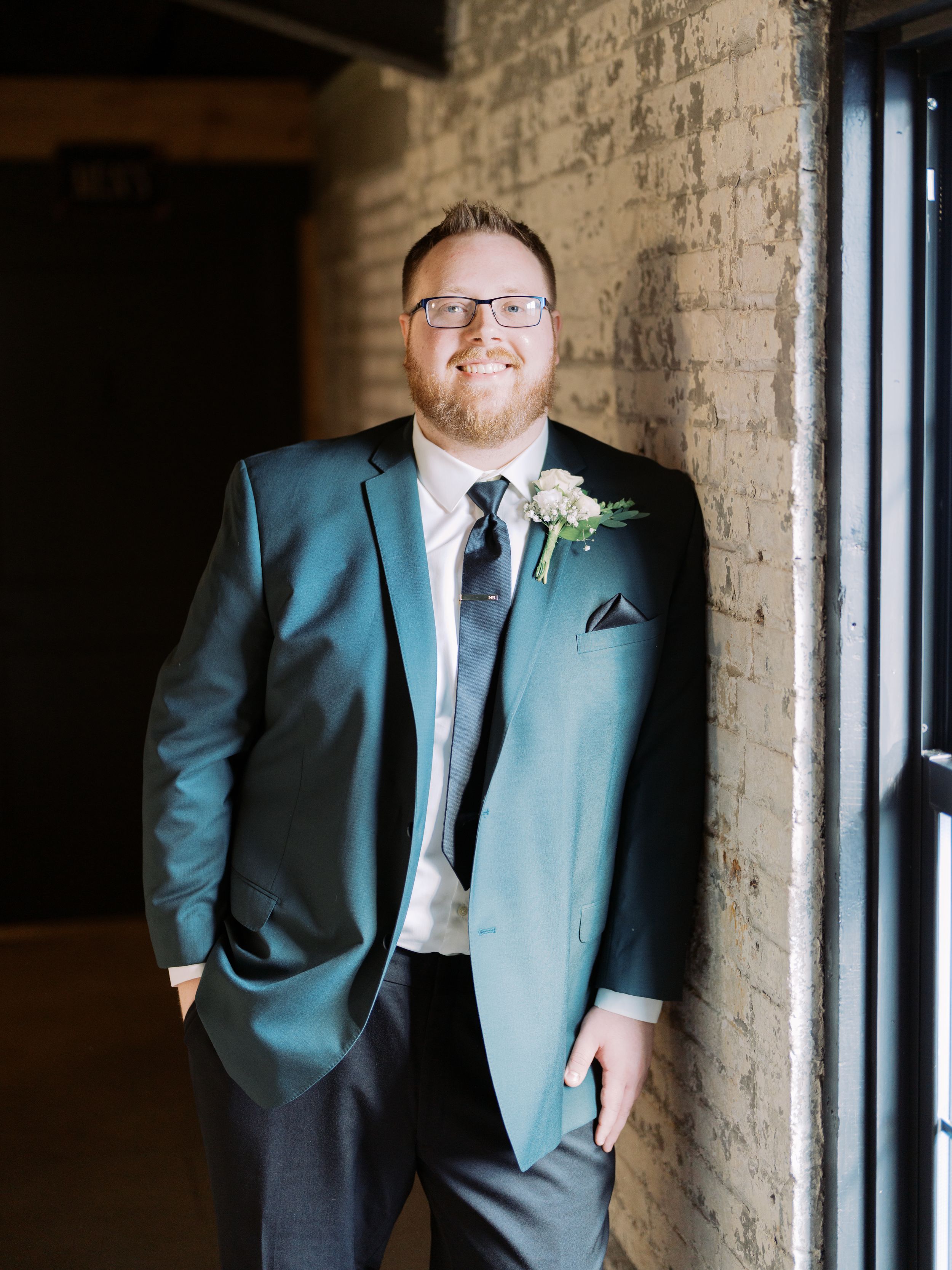 A groom in a teal suit poses against a rustic brick wall in a series of candid wedding portraits.
