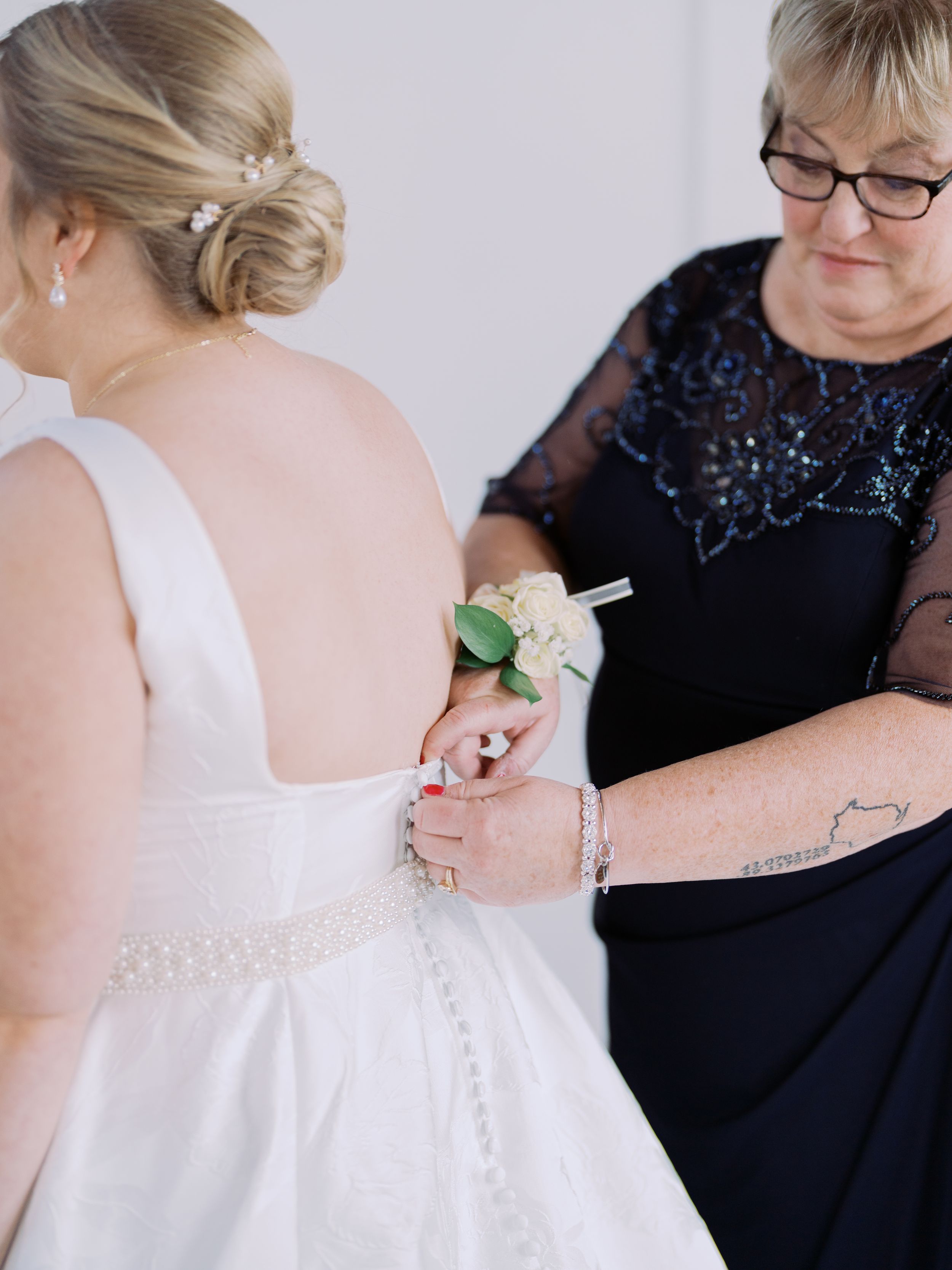 A woman in a white wedding dress being helped with buttons by another person wearing a black formal dress with sparkles.