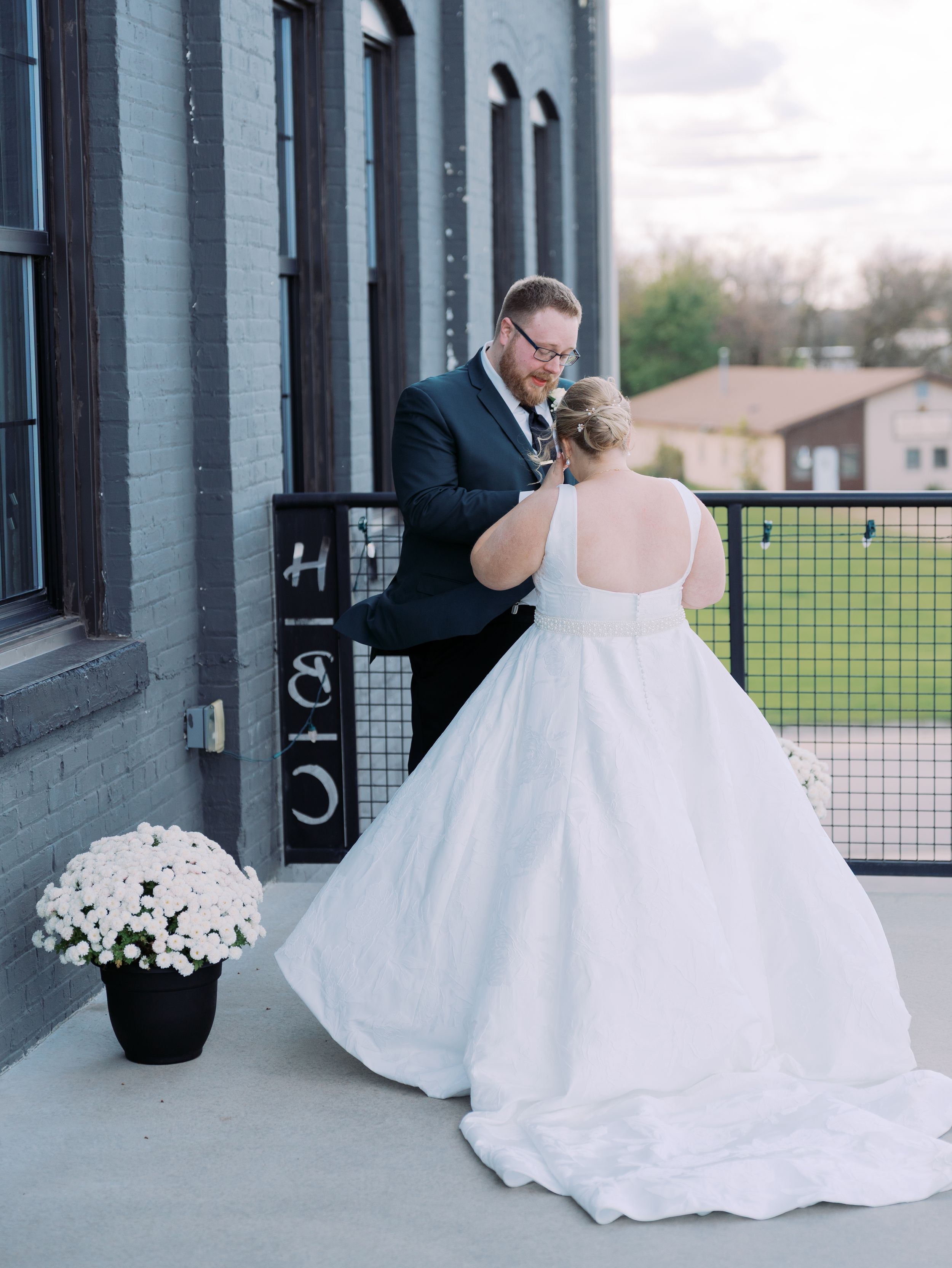 A bride in a flowing white ballgown poses elegantly in an industrial-style venue with large windows and green fields outside.