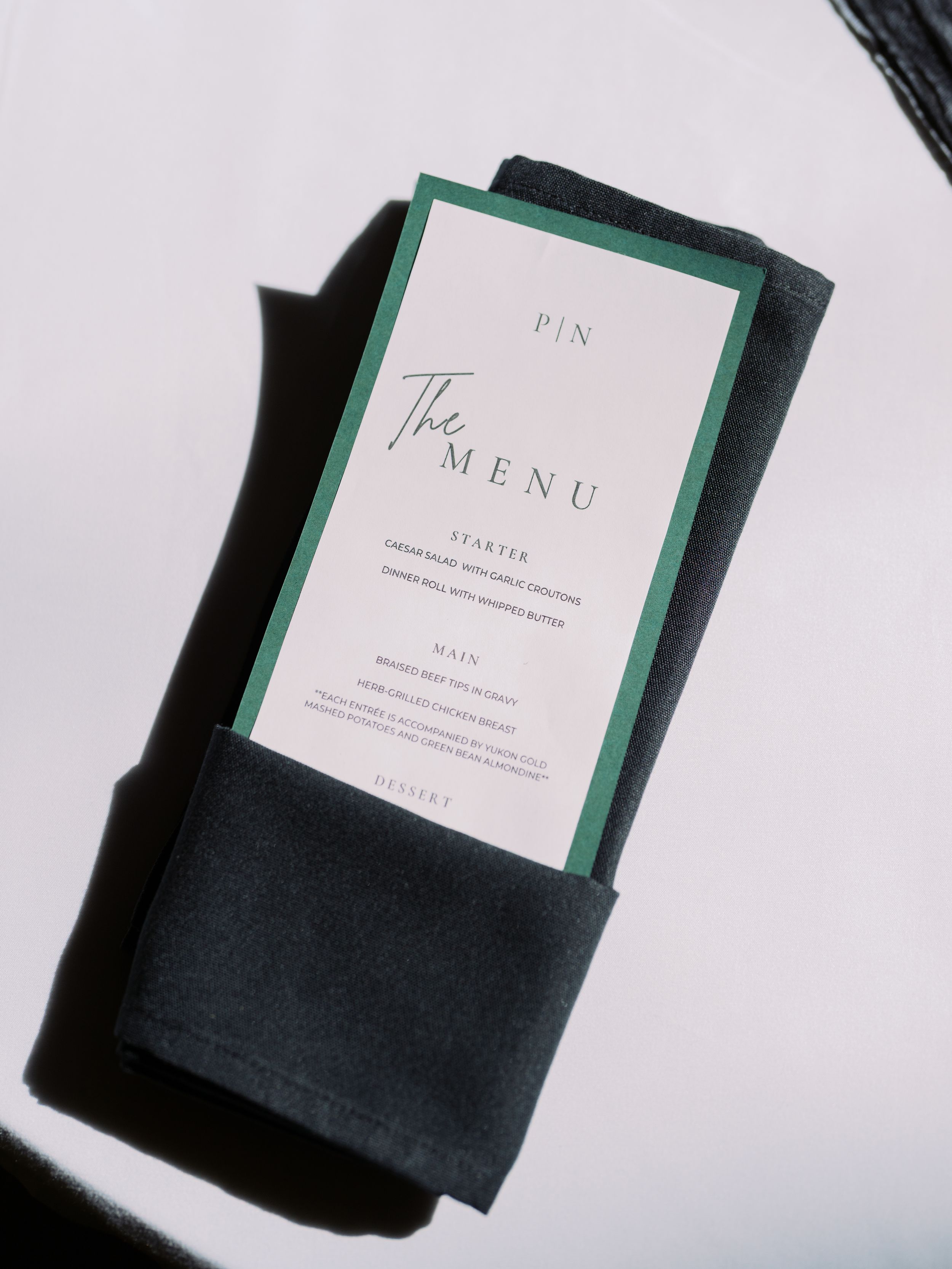 An elegant restaurant menu card with green border trim tucked into a black napkin on a white surface.