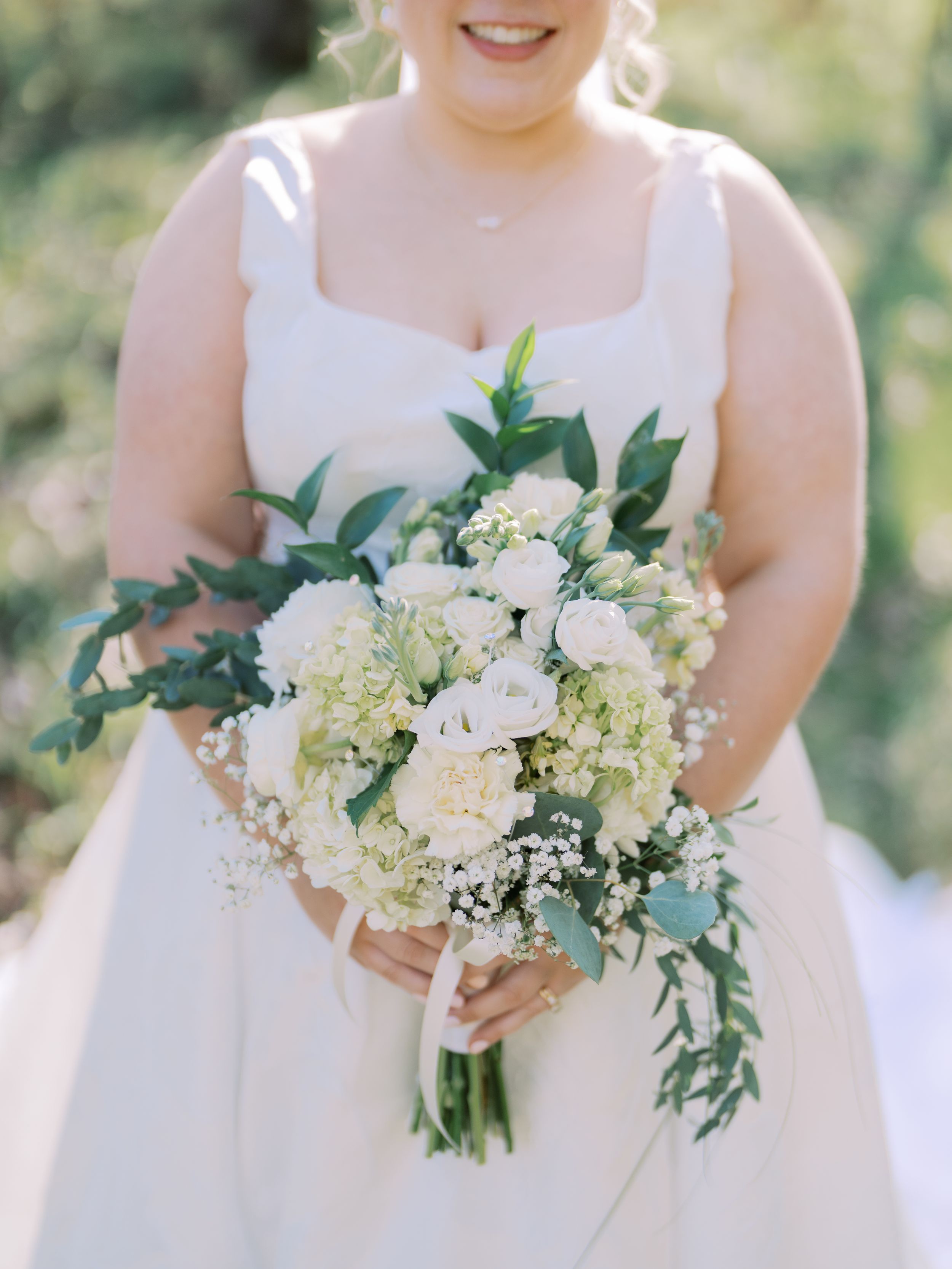 A wedding bouquet with white roses, hydrangeas and greenery held against a white dress in natural lighting.