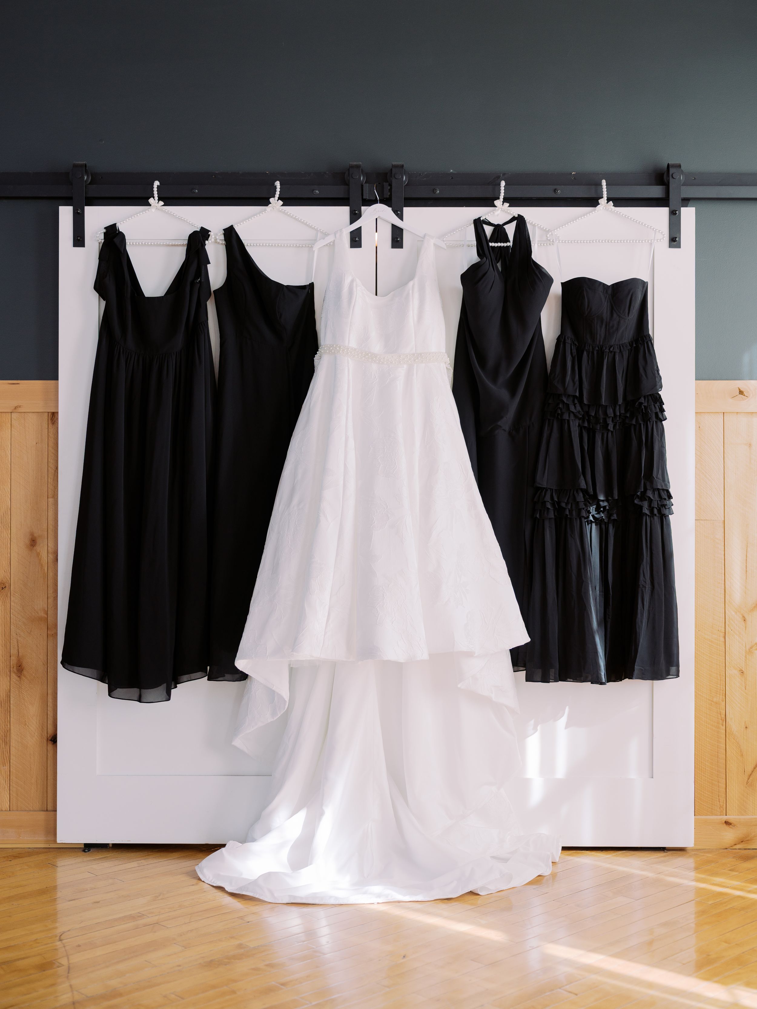 A collection of black bridesmaid dresses and a white wedding gown hanging on display in a room with wooden floors.