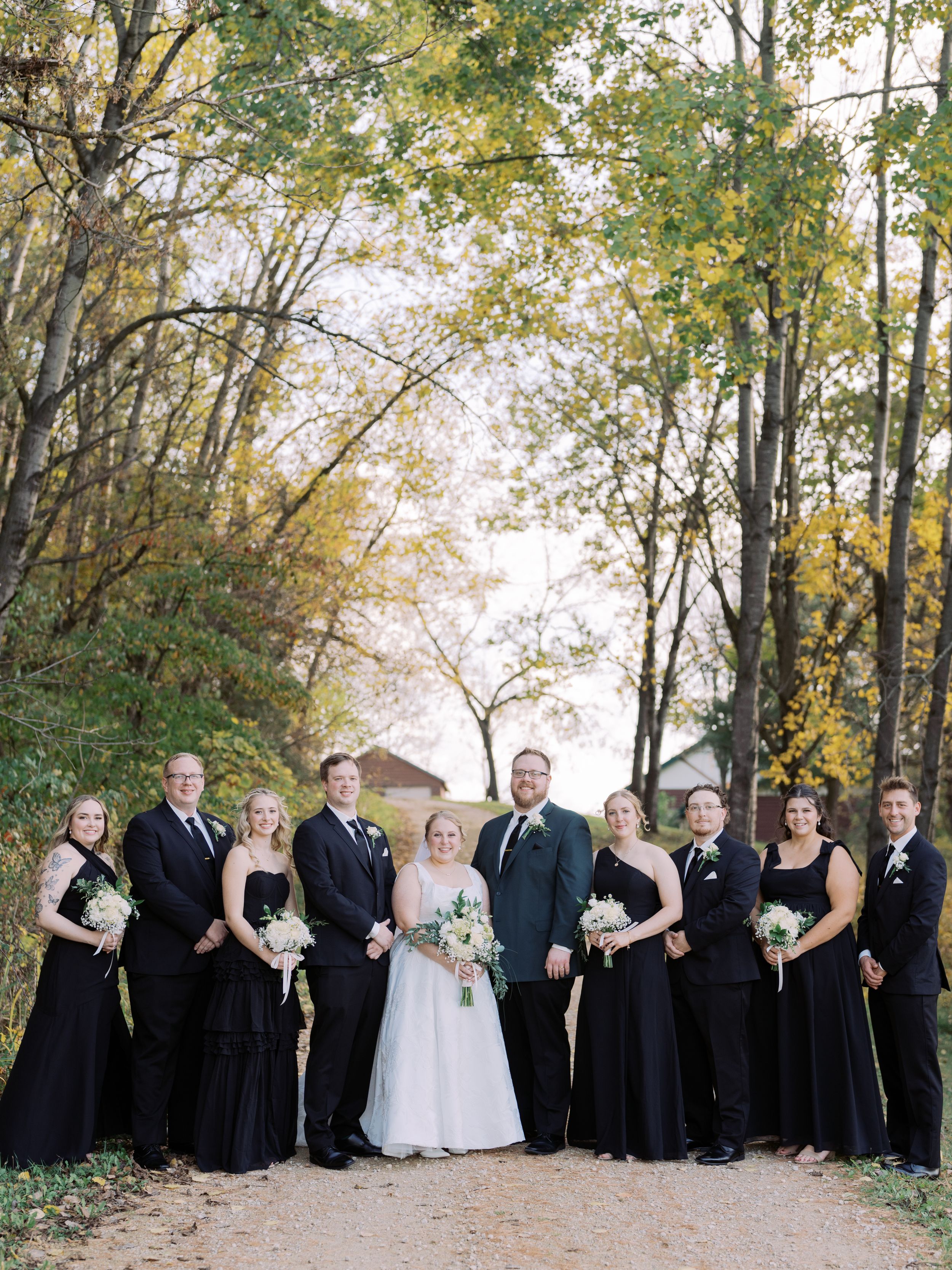 Wedding party in black and white attire poses on a tree-lined path during autumn.