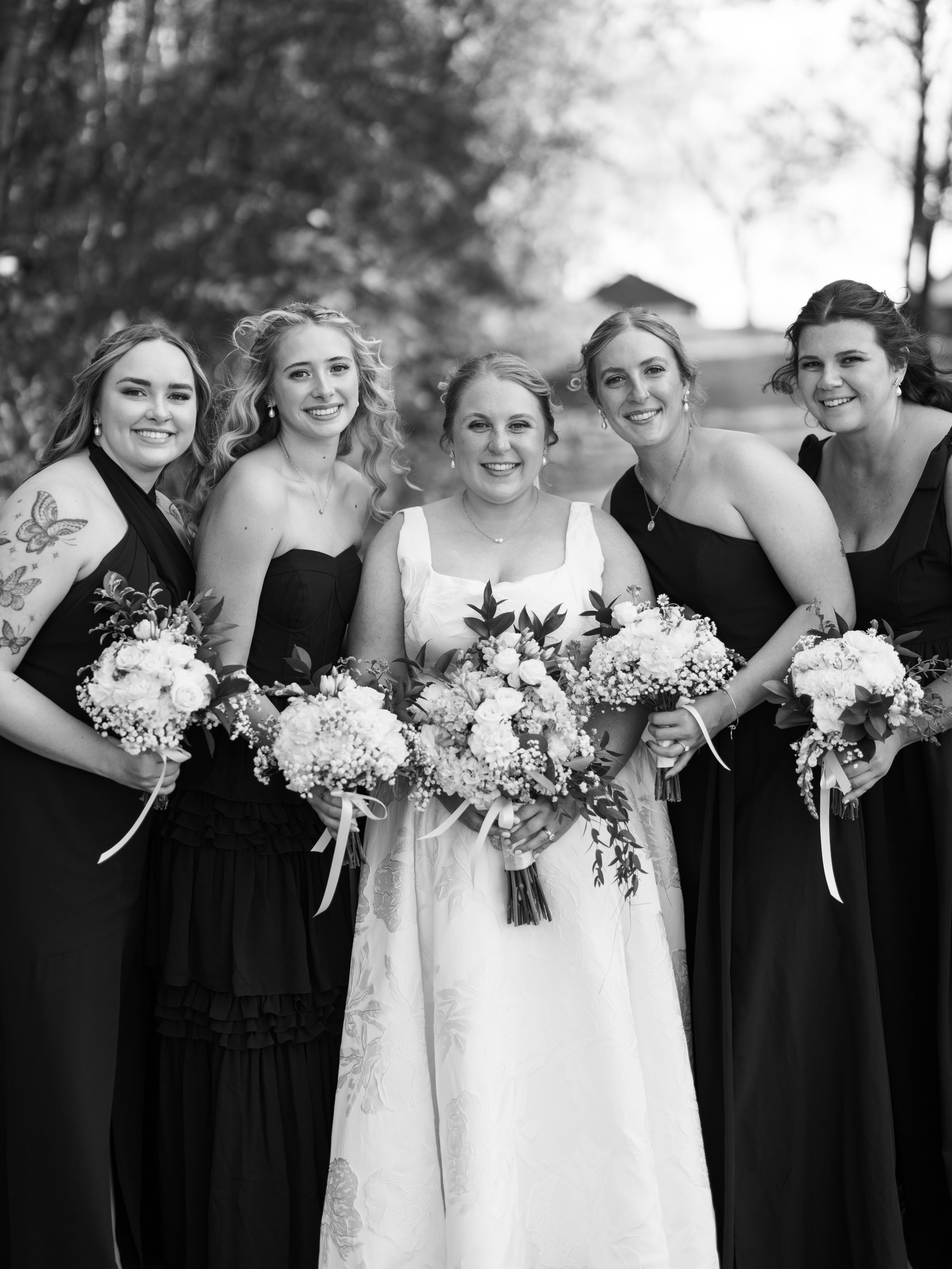 Black and white wedding party photo showing bridesmaids in dark dresses holding white floral bouquets beside bride in white.