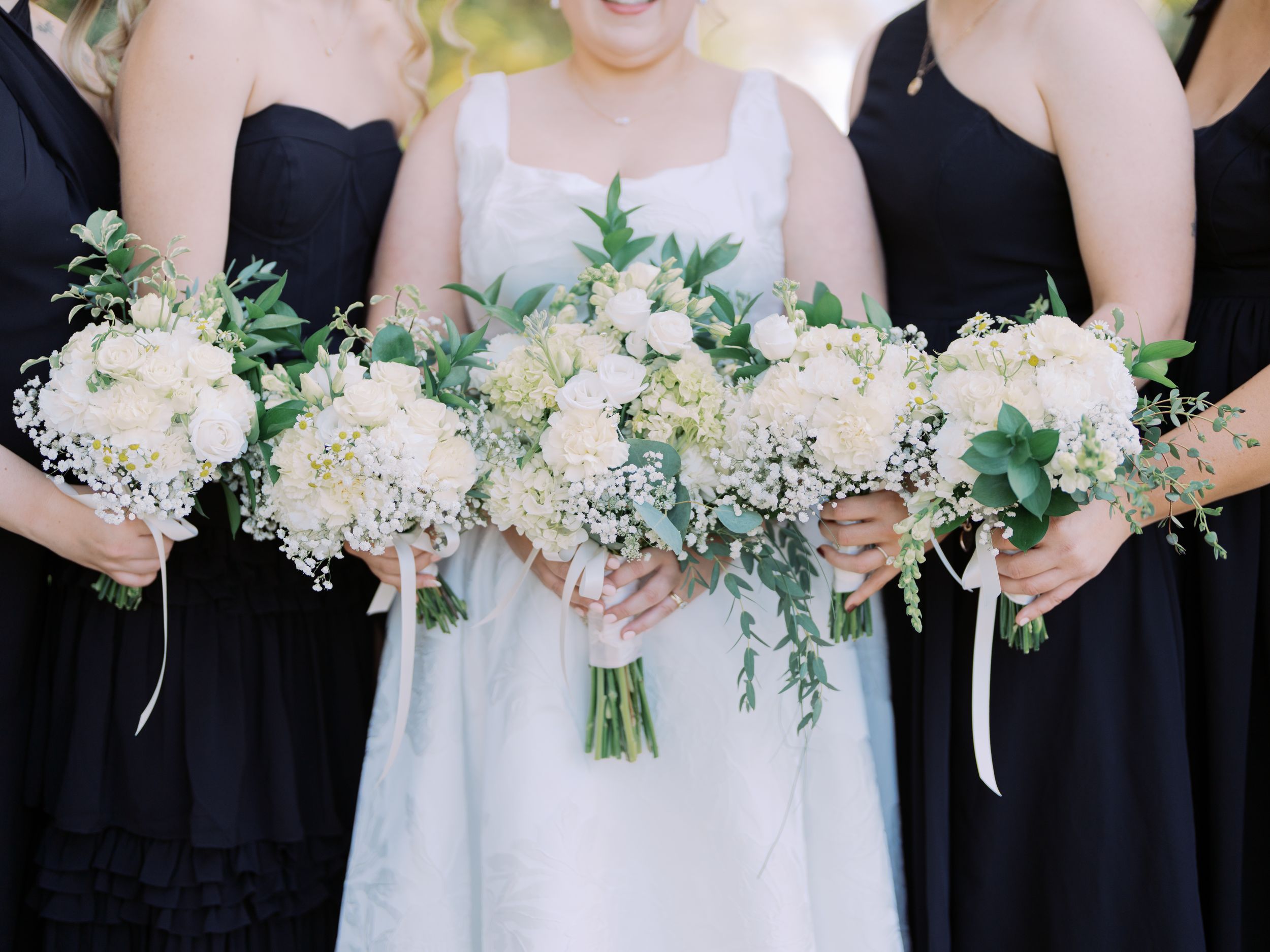 Row of bridesmaids in black dresses holding delicate white and pink floral bouquets against a light background.