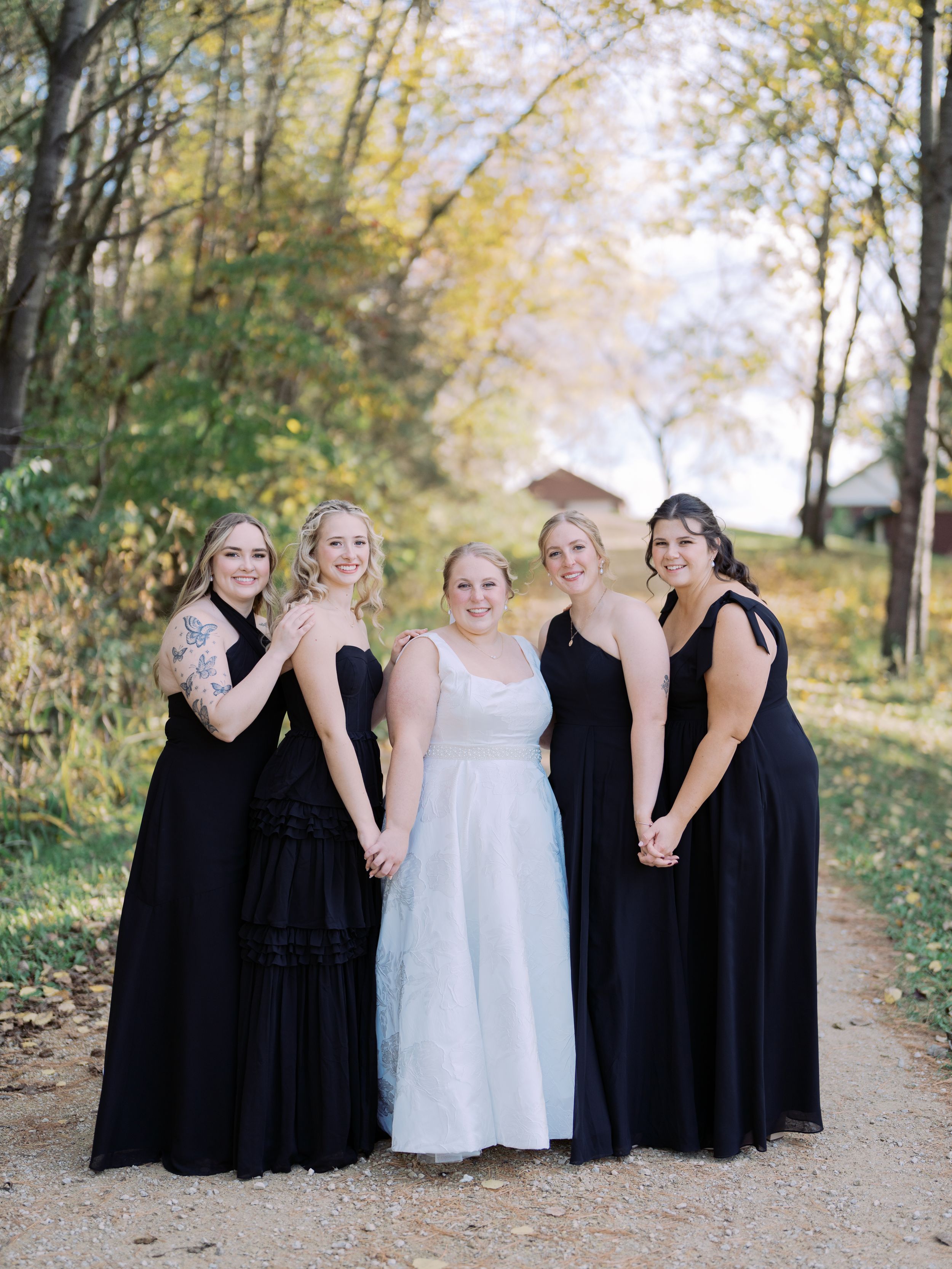 A bridal party wearing black dresses and one white dress pose together on a tree-lined path during autumn.