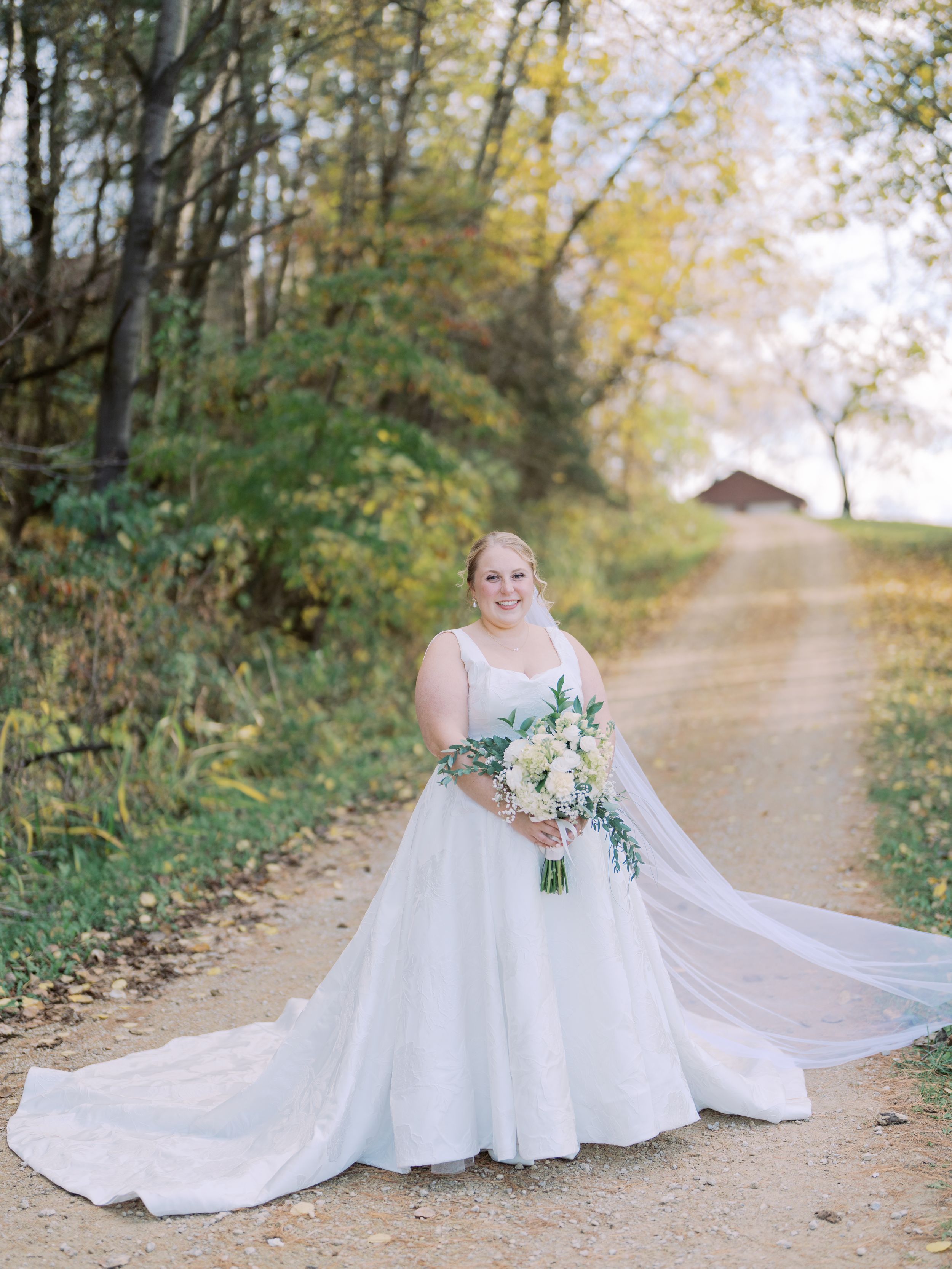 A bride in a flowing white wedding gown stands on a sunlit autumn path surrounded by golden trees.