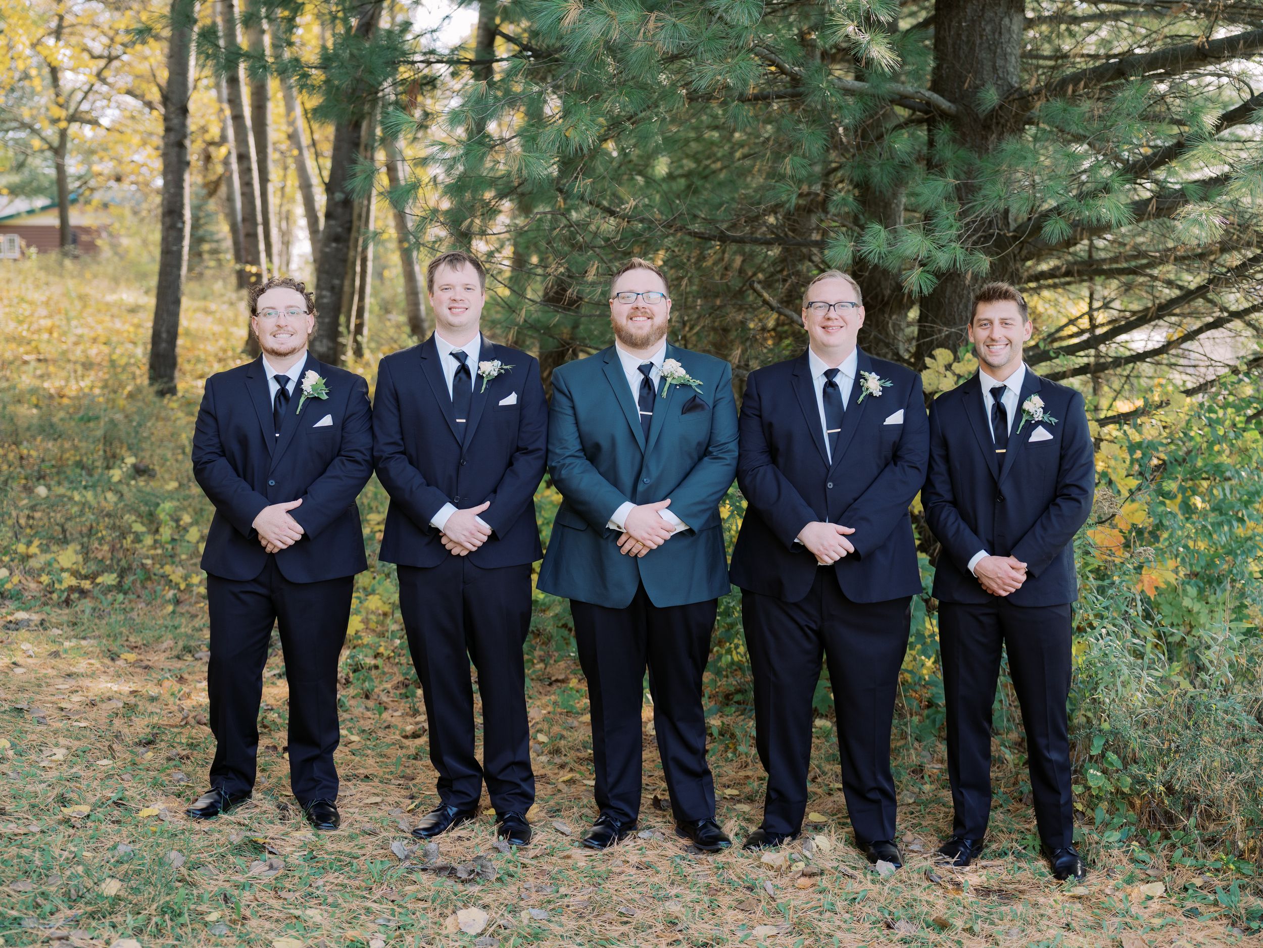 Five groomsmen in dark suits stand together outdoors against an autumn forest backdrop.