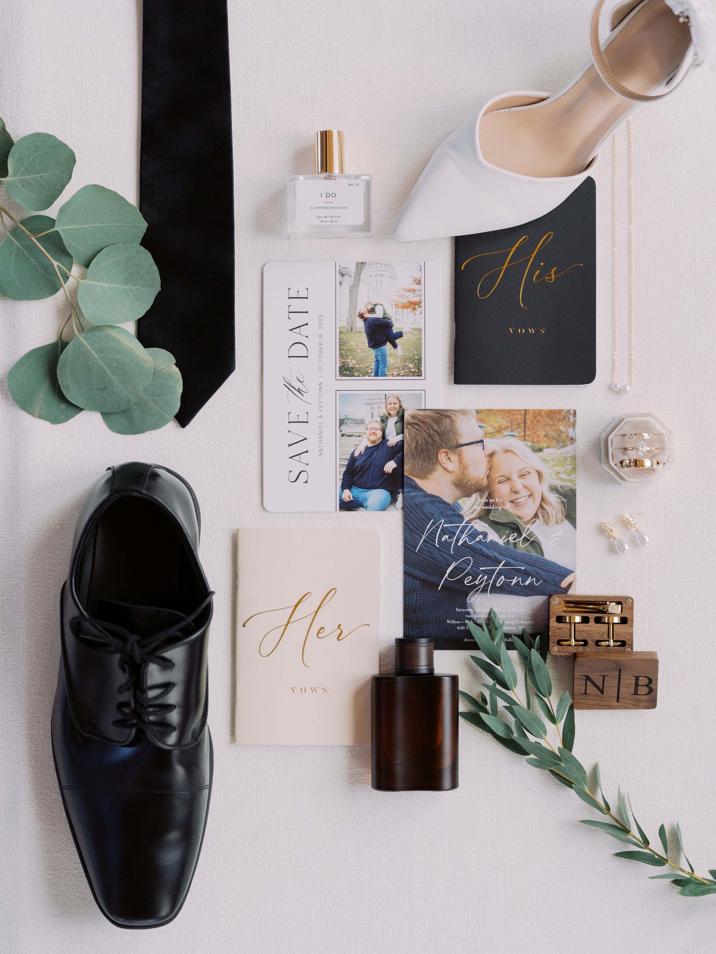 Wedding details flatlay with shoes, perfume, save the date cards, and eucalyptus leaves on white background.