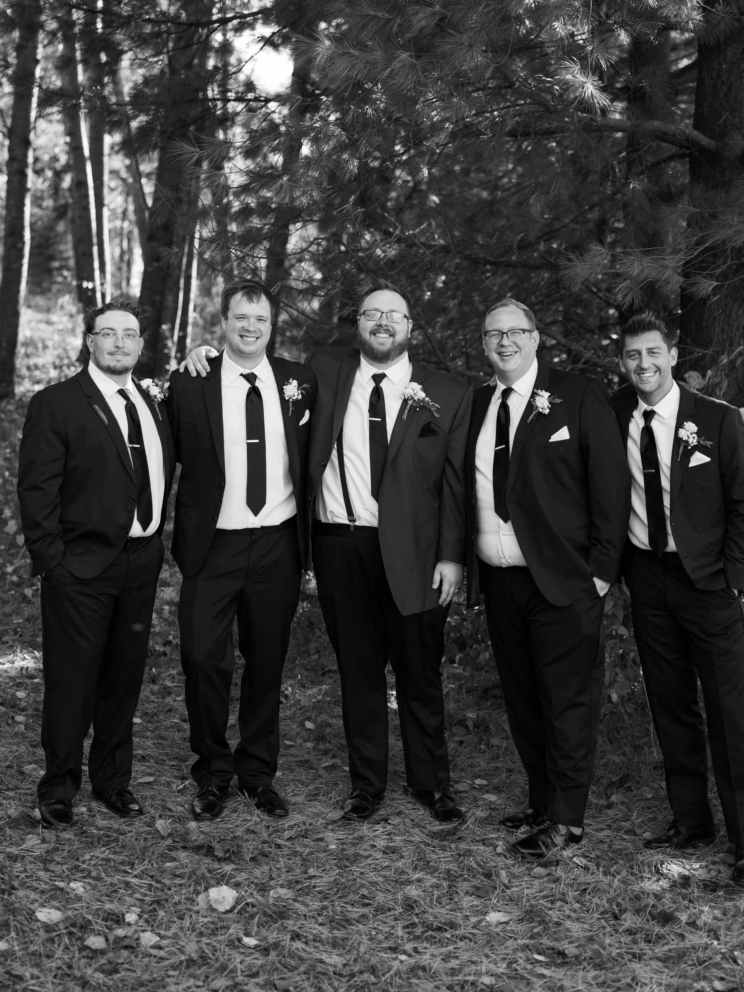 A group of groomsmen in black suits and ties pose casually while leaning against trees in an outdoor forest setting.