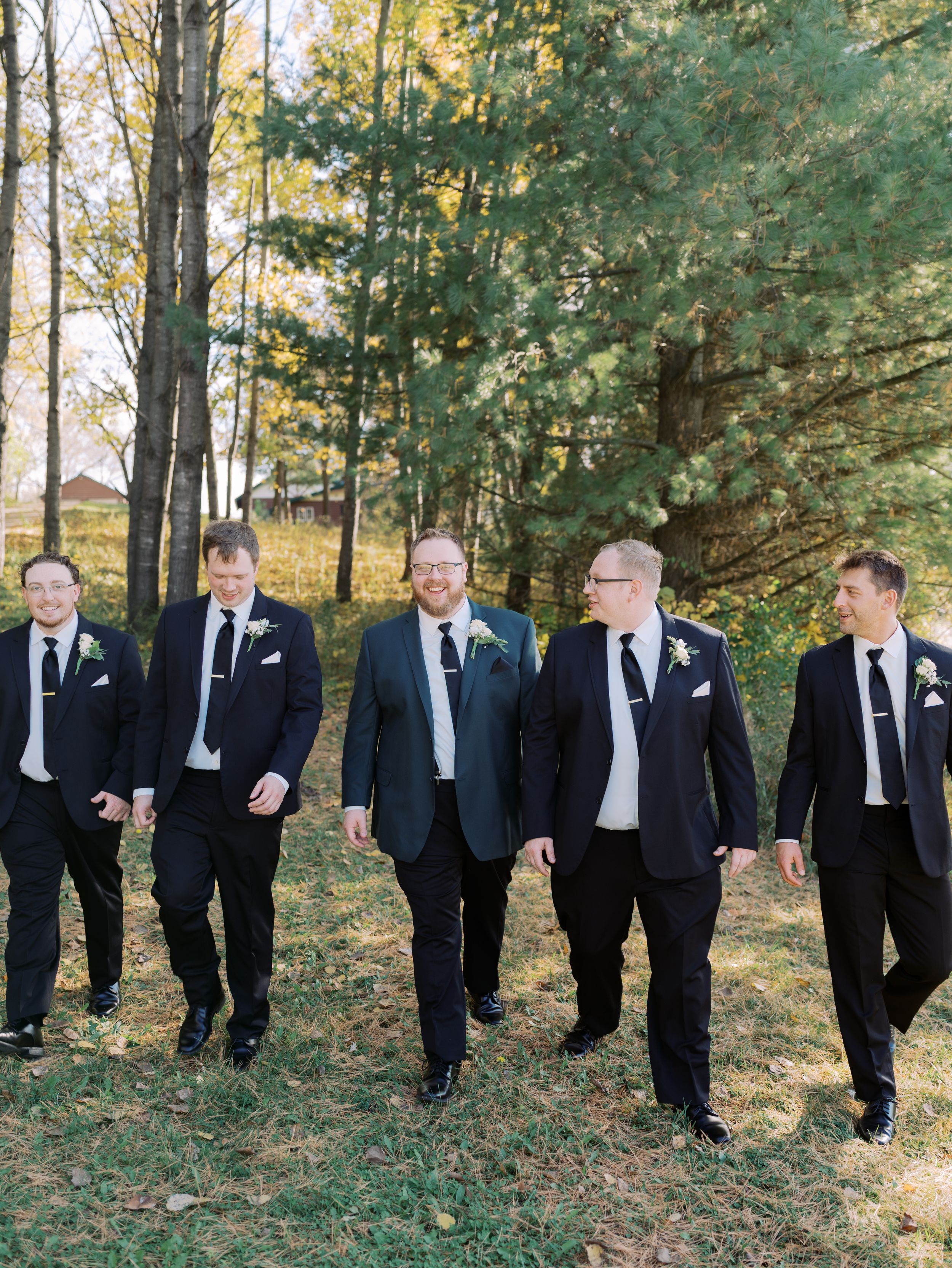 Wedding party in black tuxedos walking together through autumn trees and fallen leaves in a woodland setting.