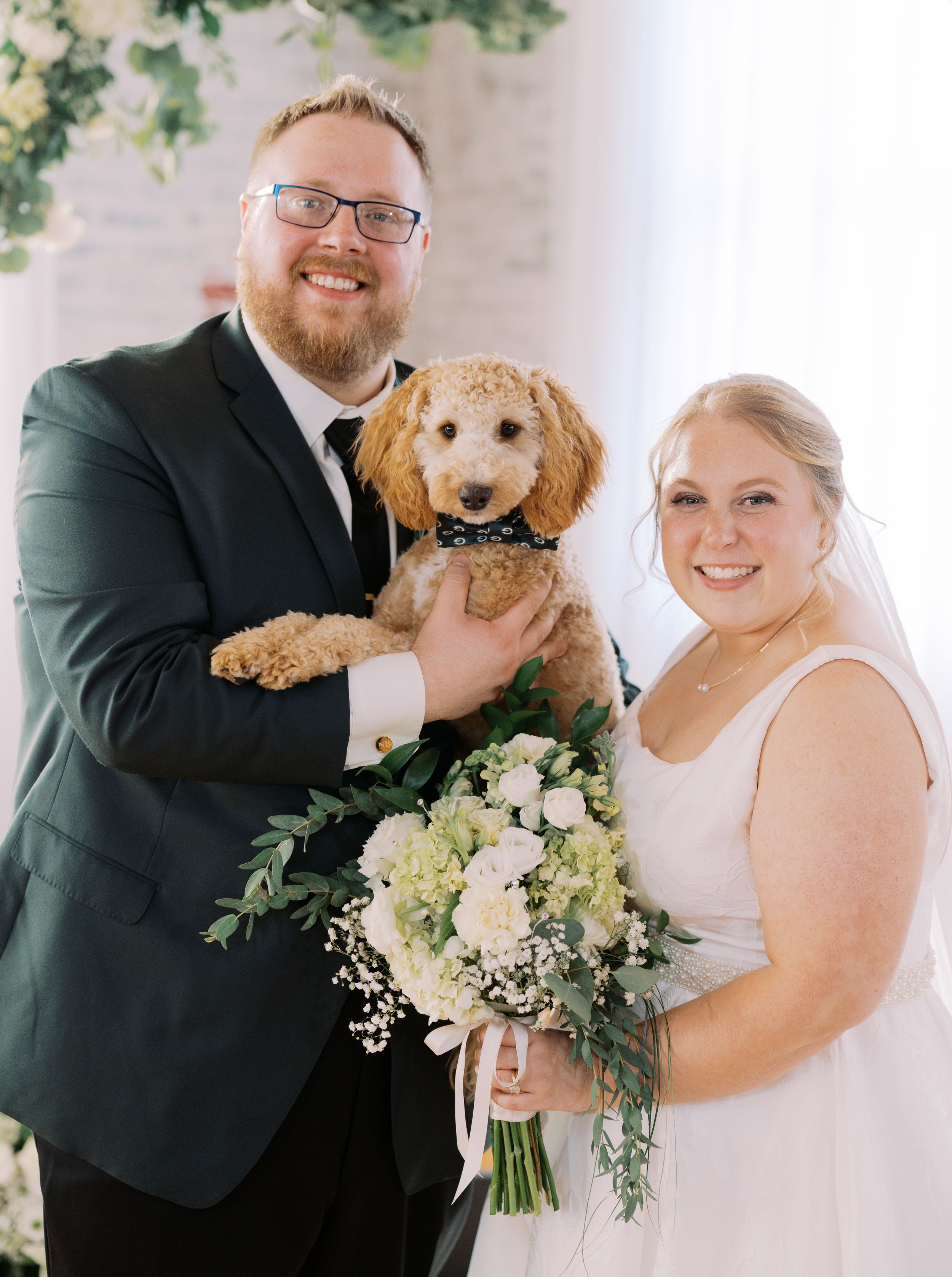 A couple in wedding attire poses with their goldendoodle dog and white floral bouquet against a light backdrop.