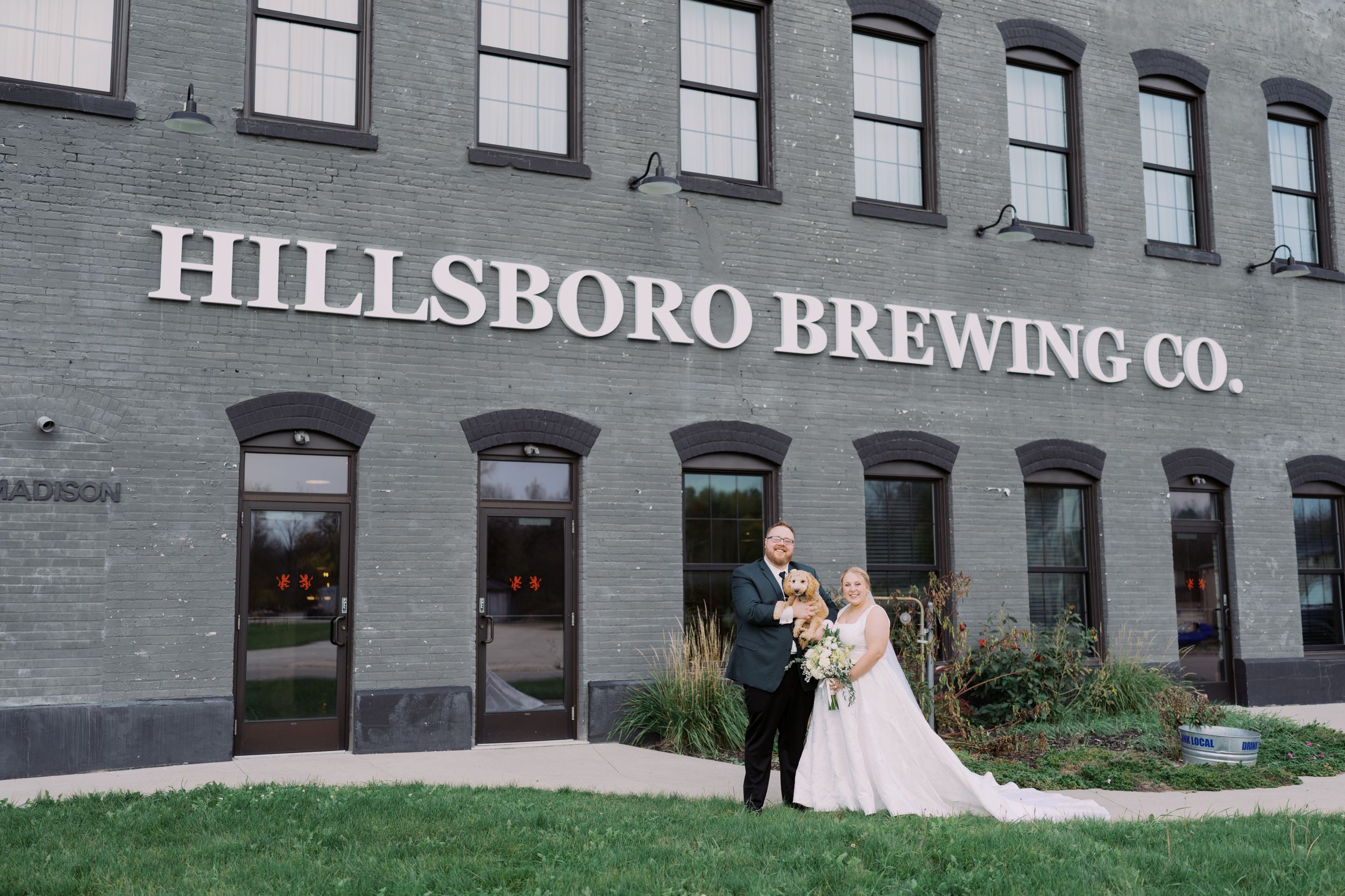 A wedding couple stands in front of the gray brick facade of Hillsboro Brewing Co. building.